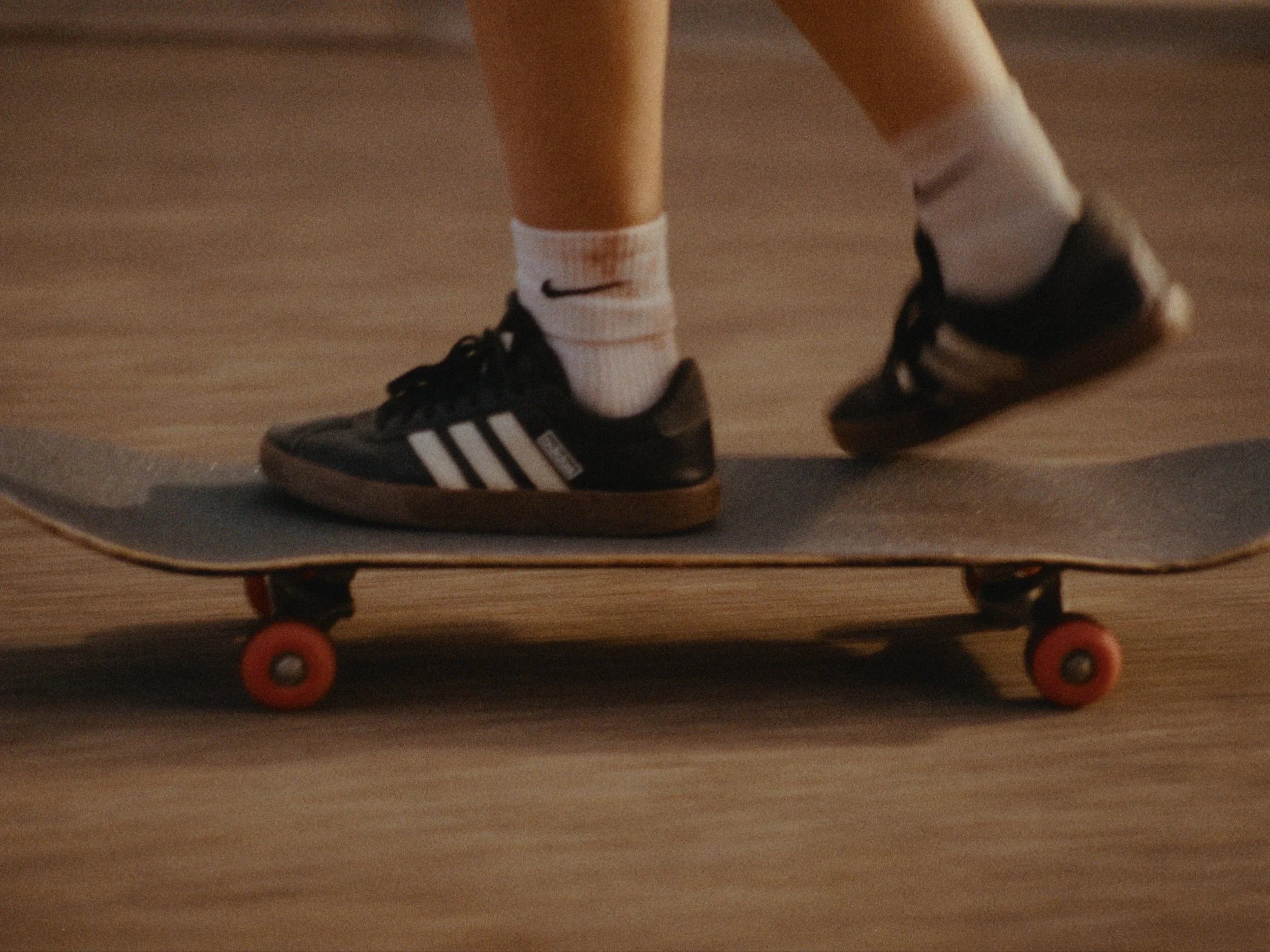 Close-up of a skateboarder’s feet wearing black Adidas sneakers and white socks, standing on a skateboard with orange wheels on a wooden floor.