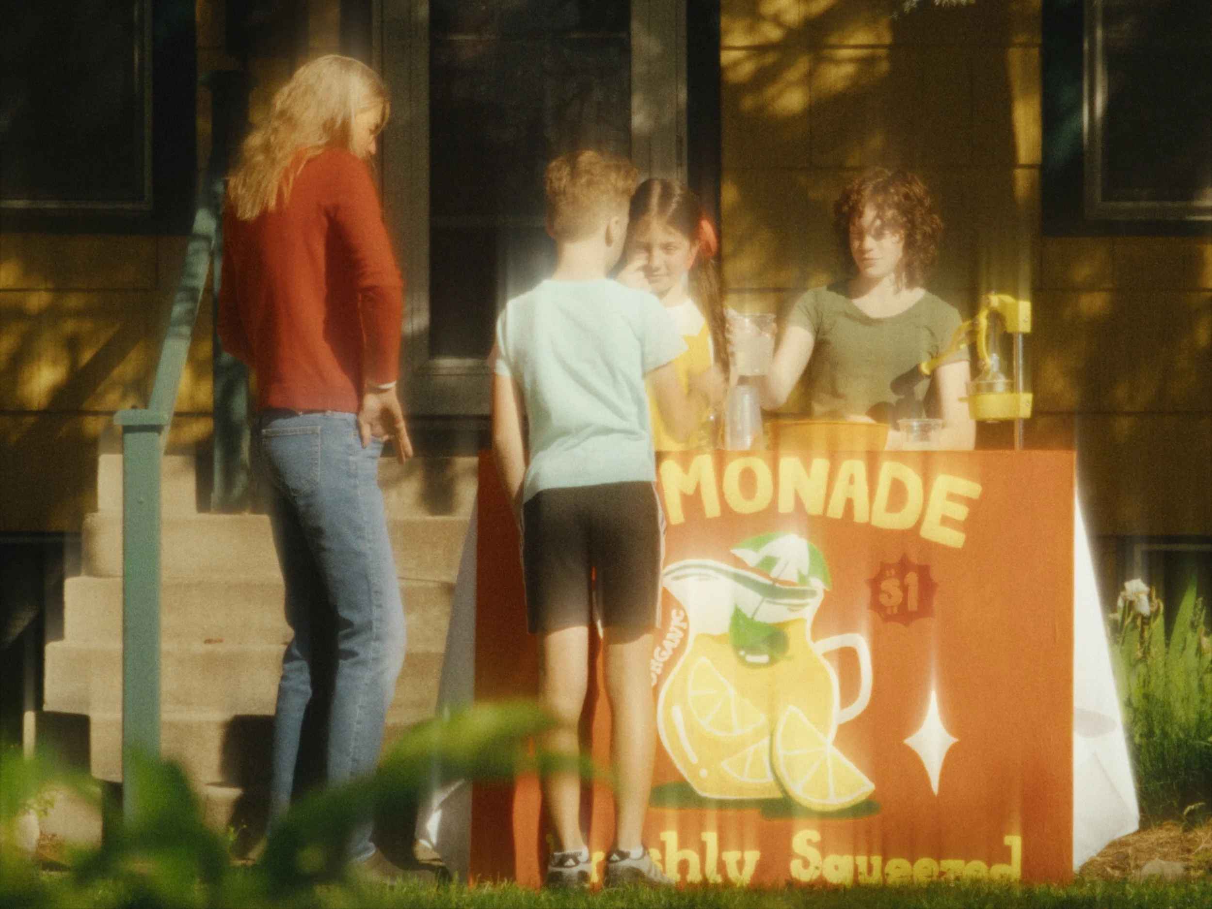 A group of children gathered around a lemonade stand with a sign that says "Lemonade" and features lemon illustrations, during evening with shadows and sunlight filtering through trees.