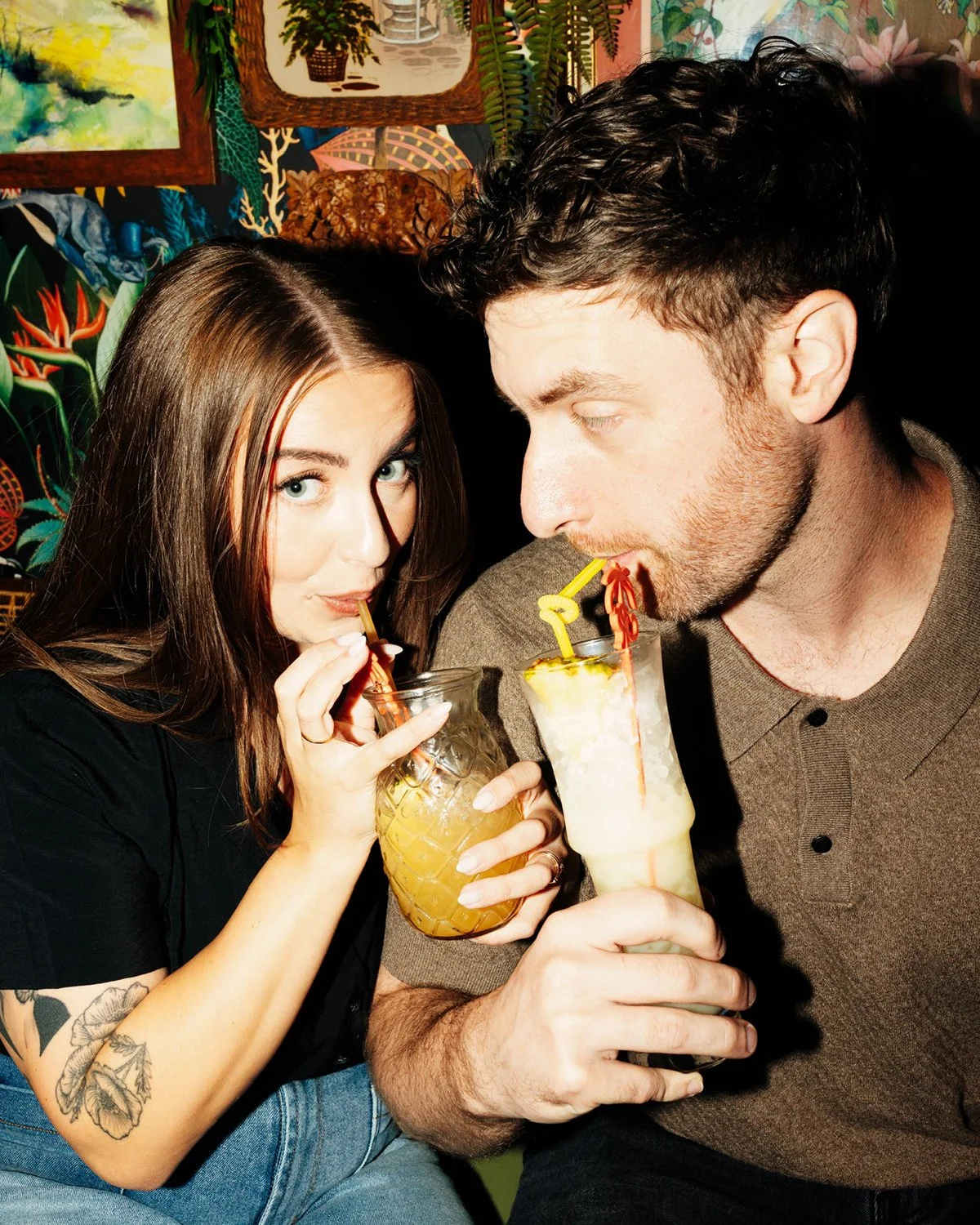 A young woman and man drinking cocktails with colorful straws inside a vibrant, tropical-themed room.