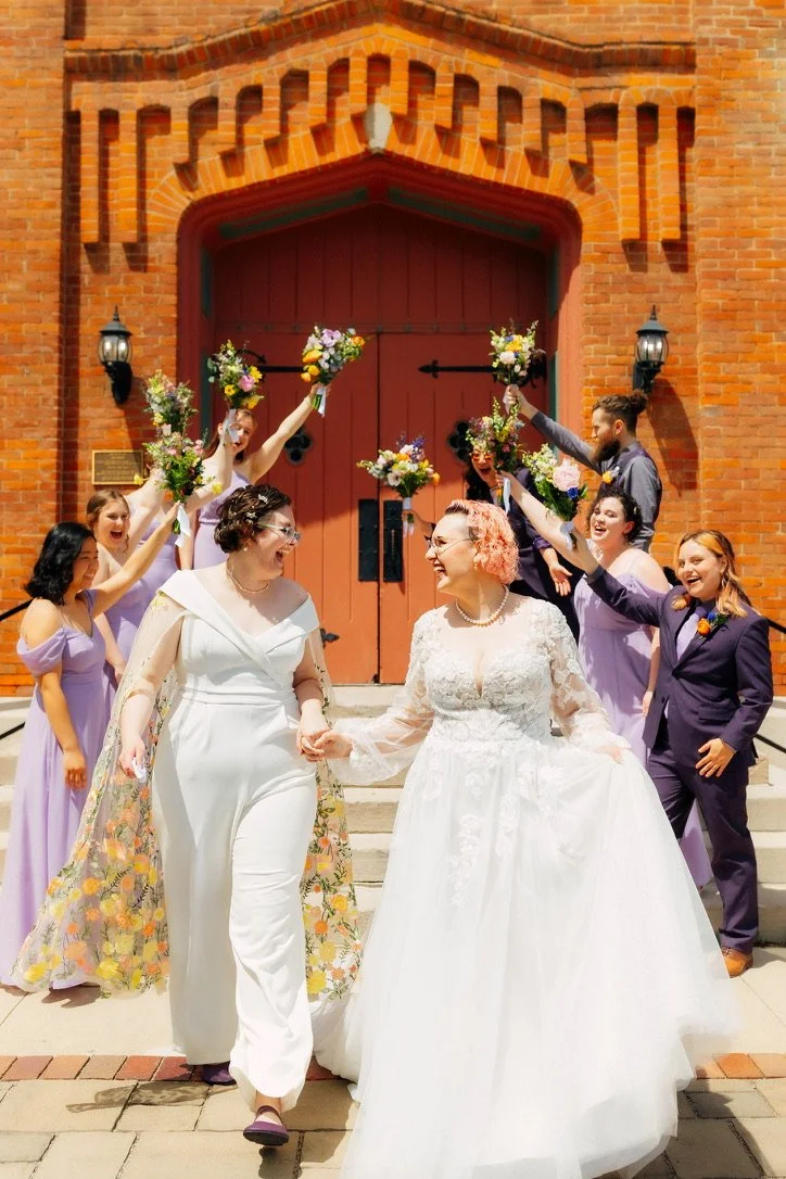 Two women in wedding dresses walking down steps after a wedding, holding hands, surrounded by friends with bouquets, in front of a brick church with red doors.