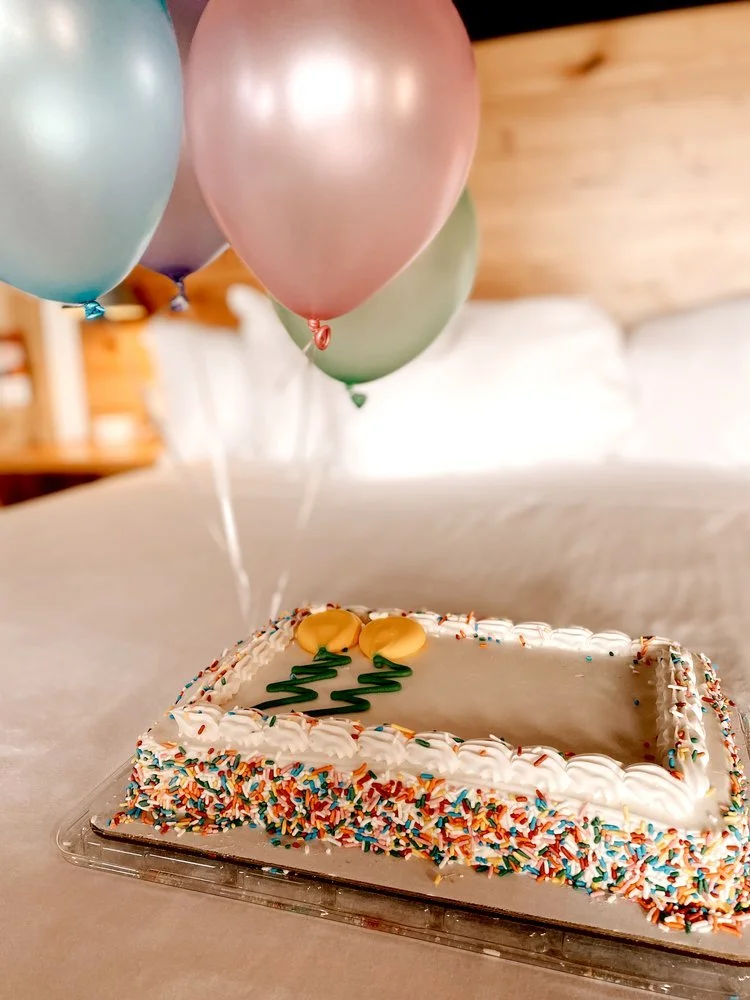 A rectangular birthday cake with colorful sprinkles and white frosting on a table, decorated with balloons floating above.