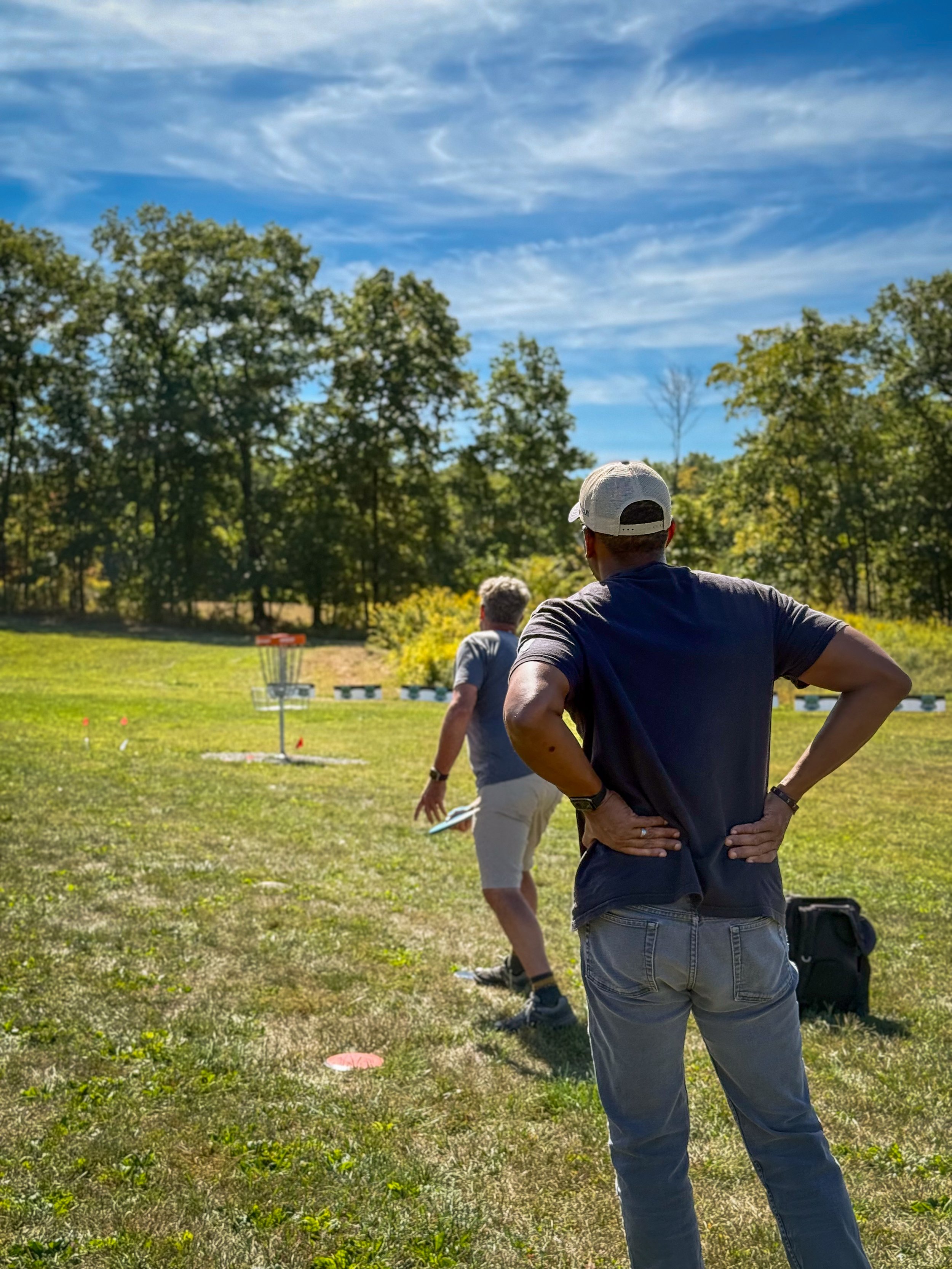 Two men playing disc golf outdoors on a sunny day, with trees and a blue sky in the background.