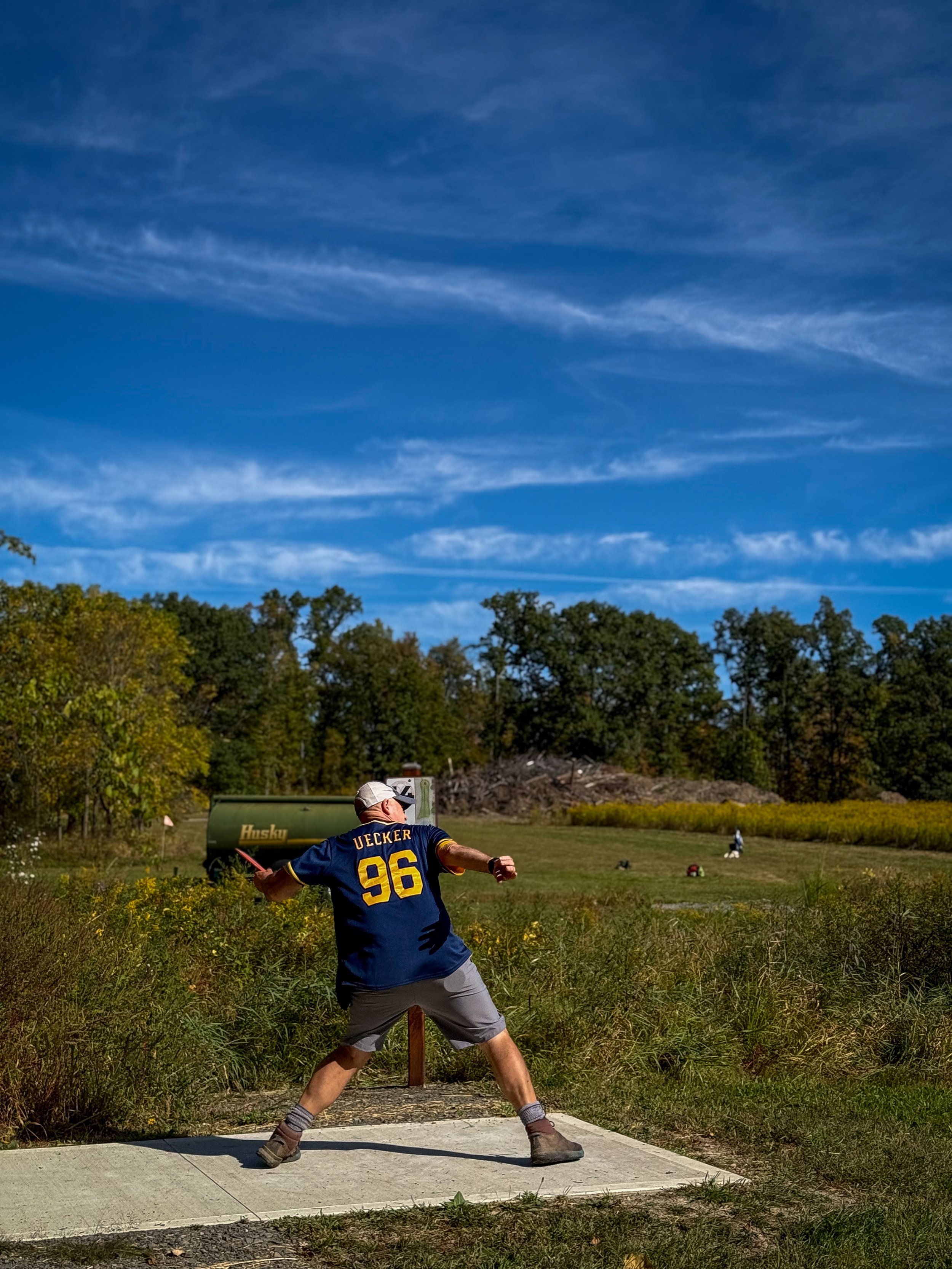 A person in a blue sports jersey with the number 96 and the name 'UECKER' is playing disc golf, throwing a disc on a grassy field with trees in the background and a blue sky above.