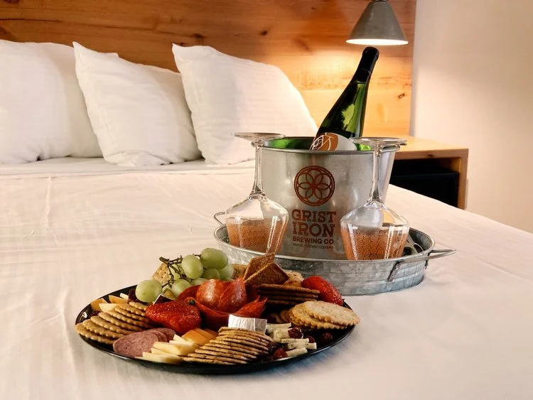 A tray with two glasses of beer, a bottle of wine in an ice bucket, and a plate of assorted crackers, cheese, grapes, strawberries, and meats on a bed with white sheets and pillows, with a wooden headboard and a lamp on the nightstand in the background.