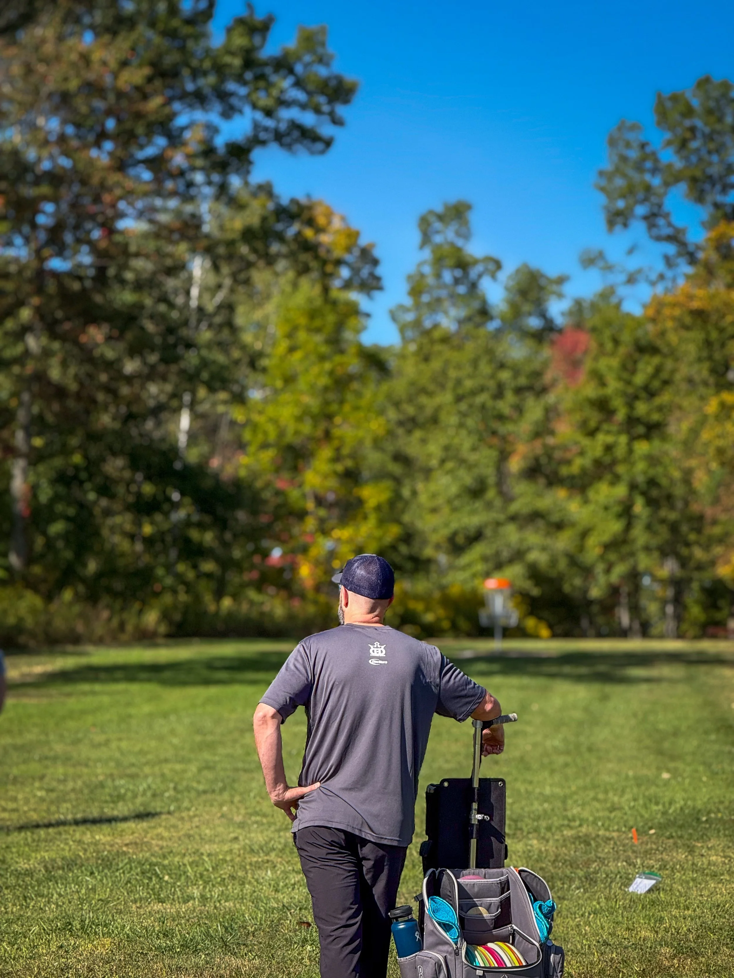 A man wearing a gray t-shirt and a black cap pushing a golf cart on a grassy field, with trees and a blue sky in the background.