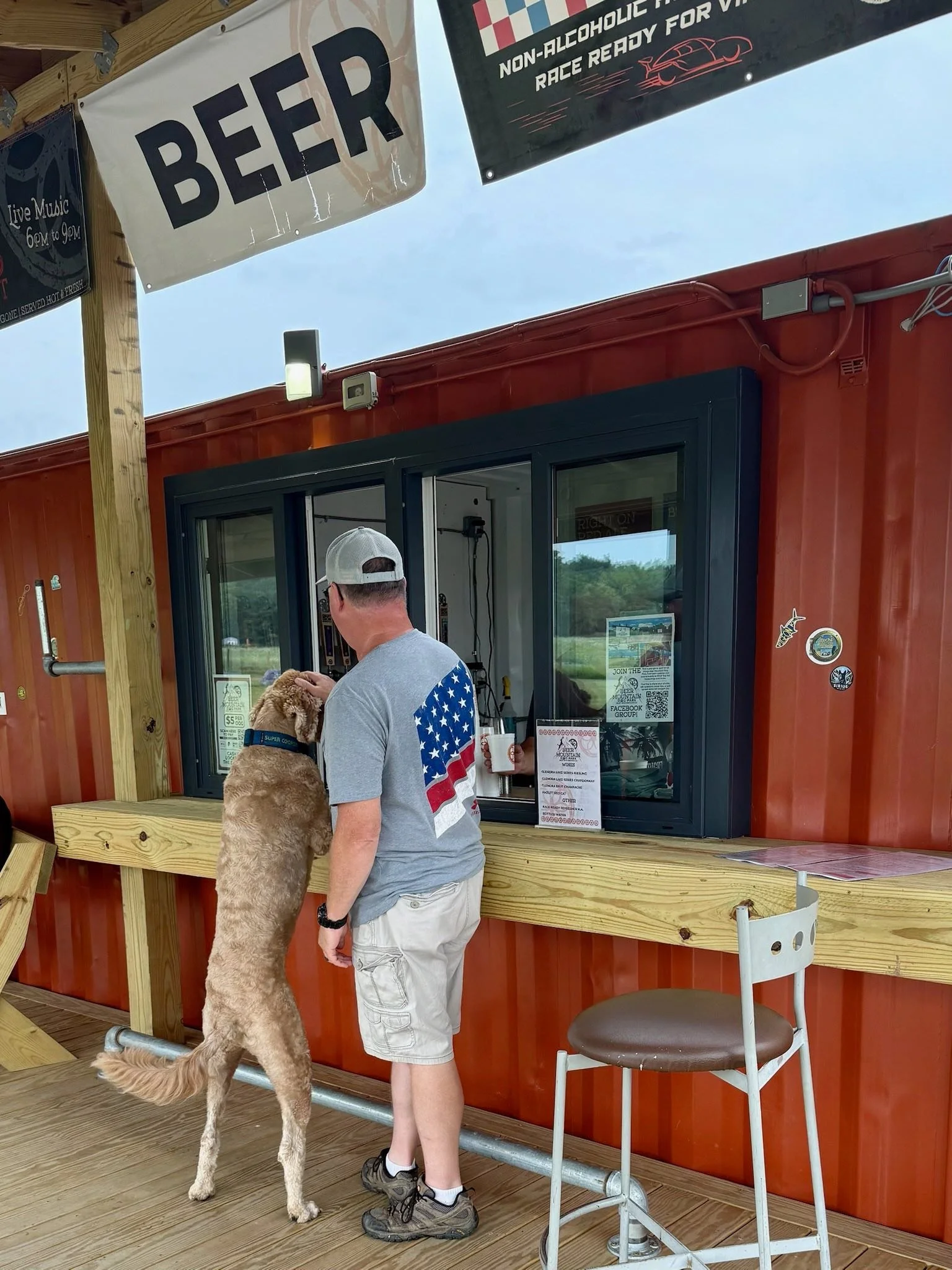 A man with a gray cap, gray T-shirt with an American flag design on the back, and cargo shorts standing at a counter with a large retriever dog at his side. The dog is standing on its hind legs leaning against the counter, interacting with the man. T