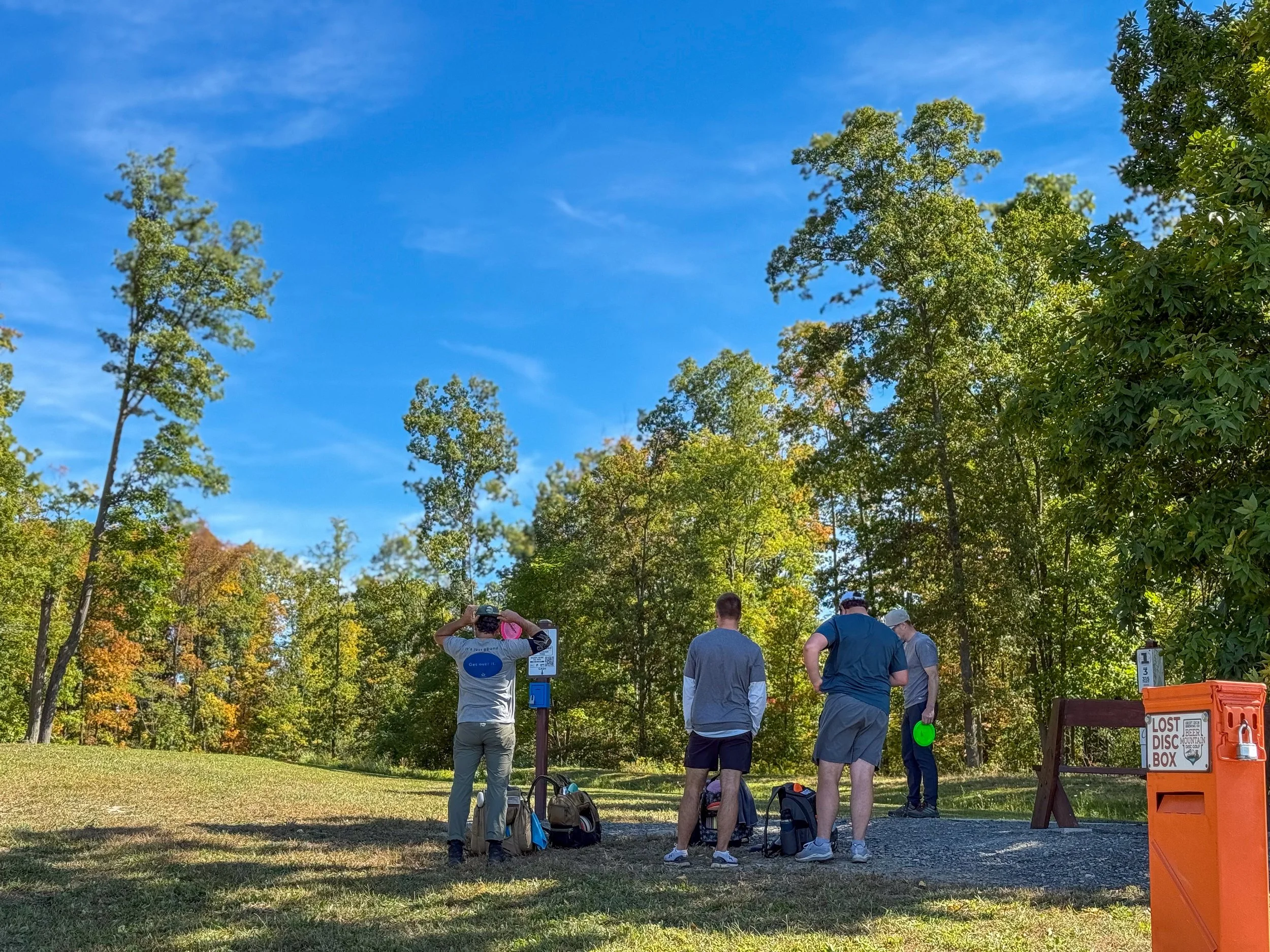 Four men standing outdoors on grass near trees, with one taking a photo and others talking or looking down, in a park-like setting under a blue sky.