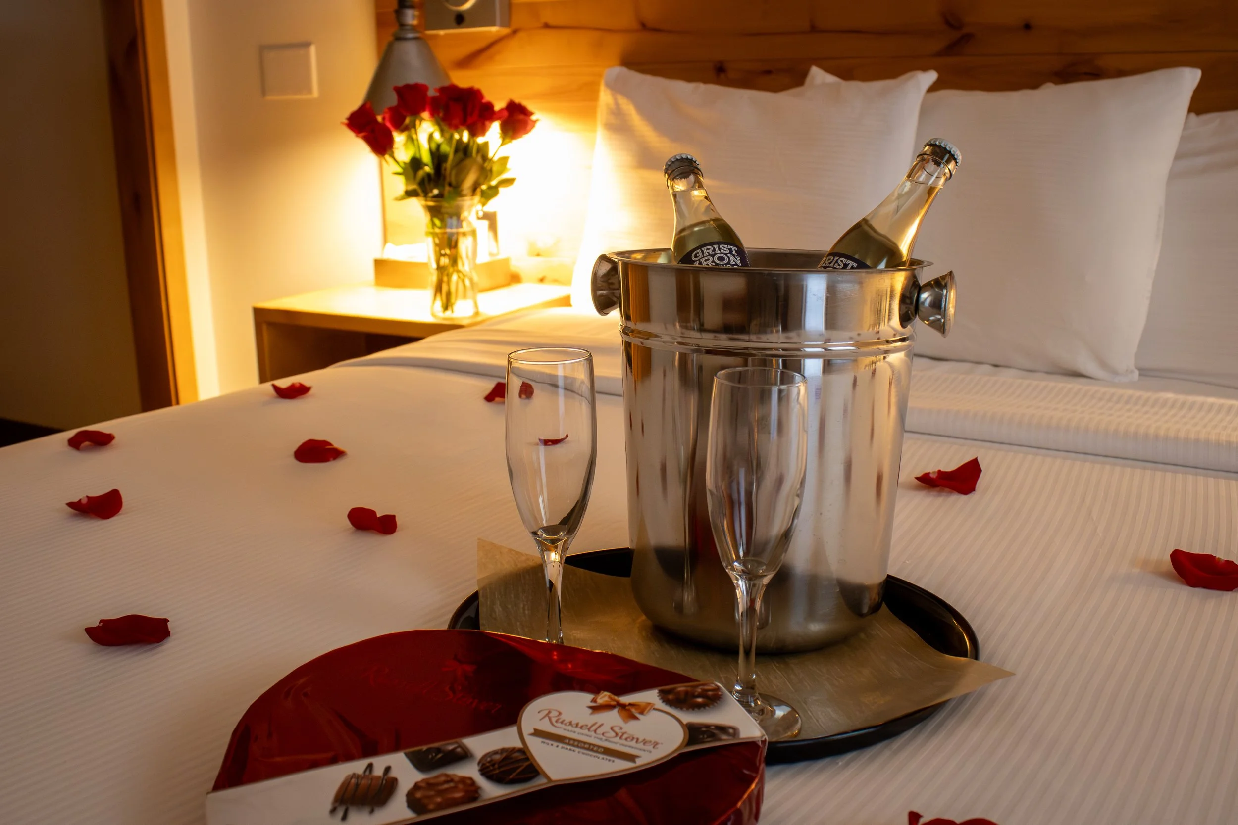 A hotel room with a bed, red rose petals scattered on the white bedspread, and a tray holding two champagne glasses, a bucket with two bottles of beer, and a box of Whitman's sampler chocolates, with a vase of red roses on a bedside table in the background.