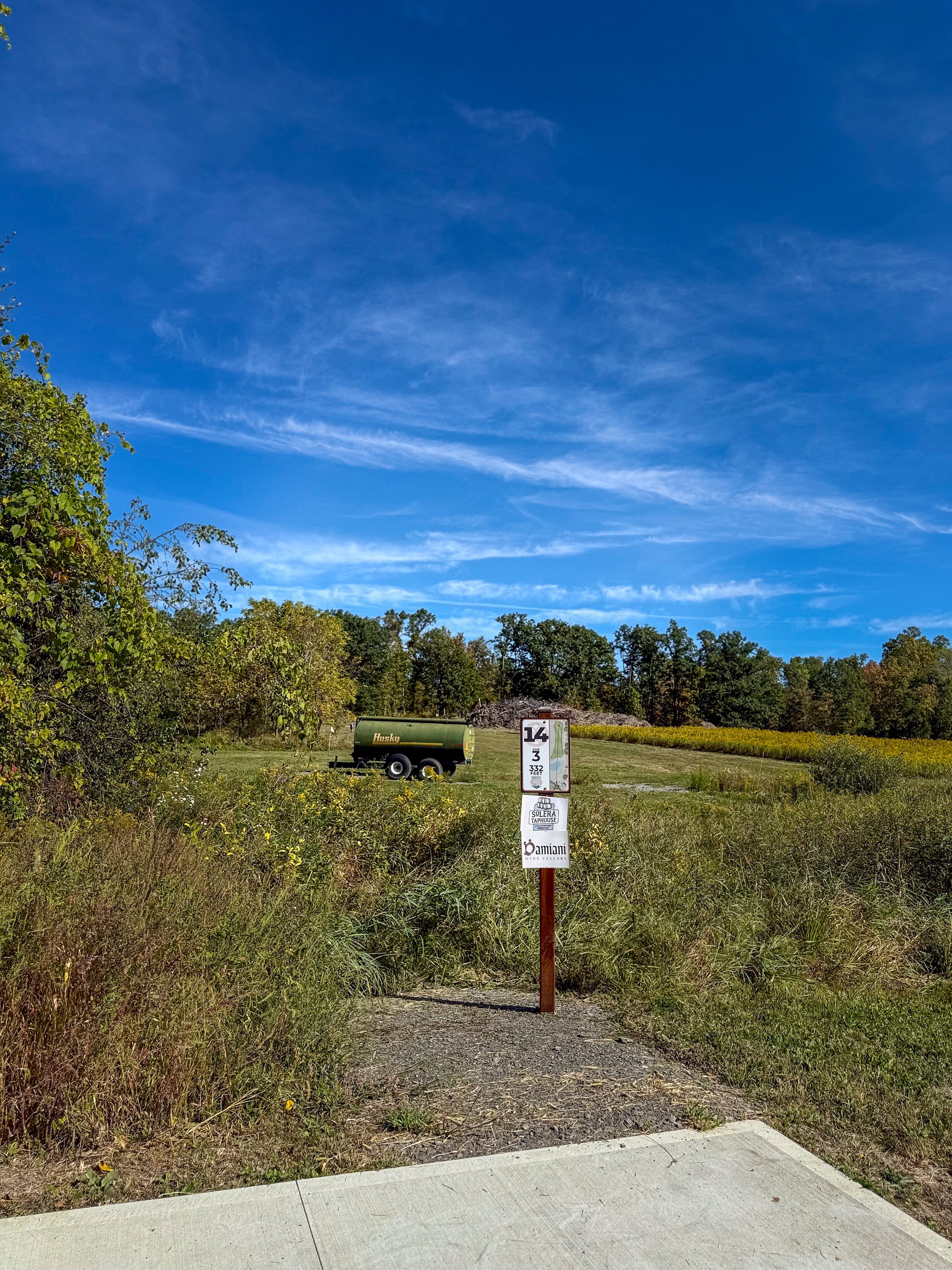 Pathway leading to a grassy field with a green water trailer in the background. There are signs on a pole in the middle of the path indicating '14 3 332' and advertisements for local businesses. Trees line the horizon with a bright blue sky and wispy