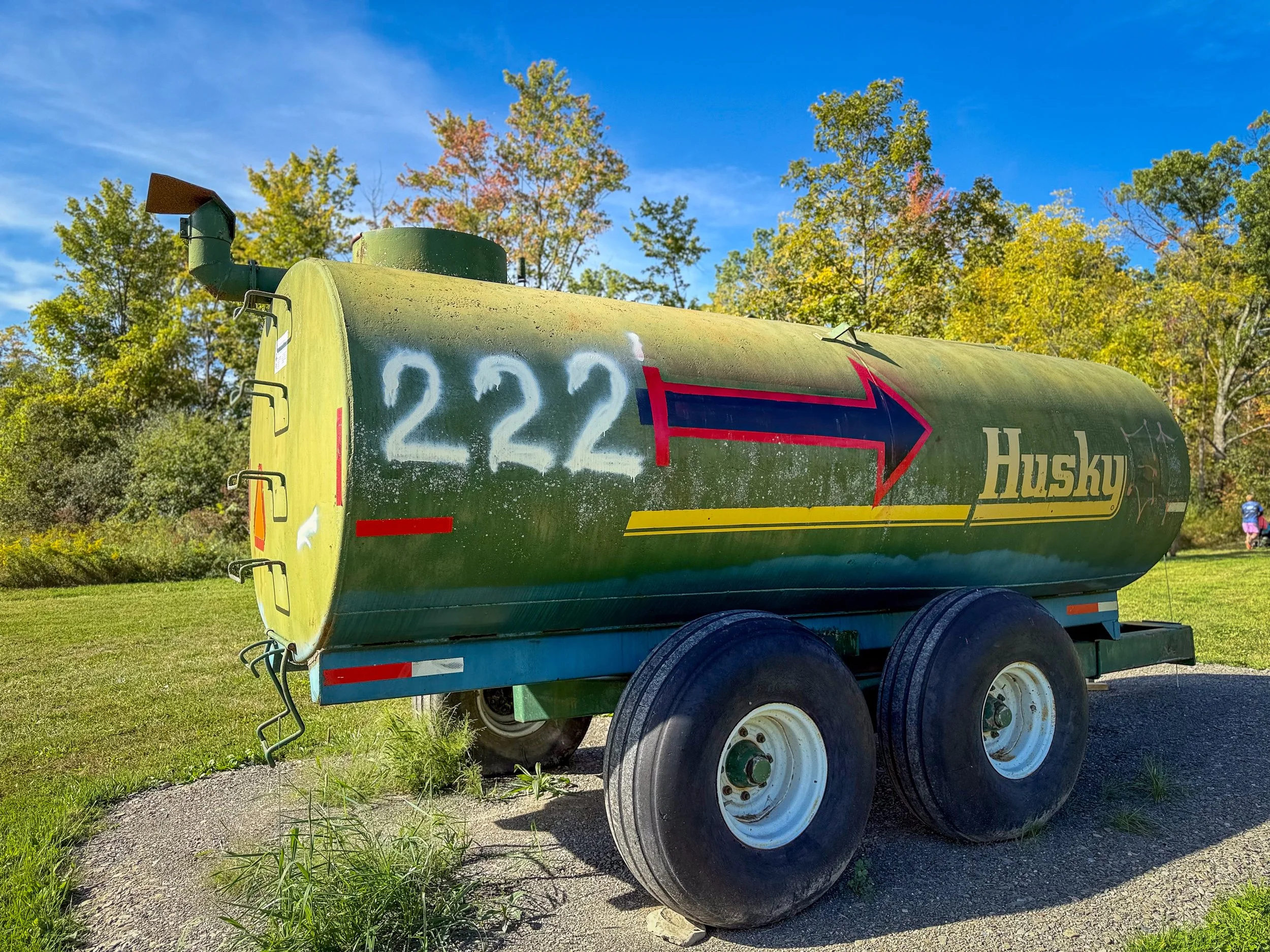 A green Husky water tanker trailer with the number 222 painted on it, parked on grass with trees and a blue sky in the background.