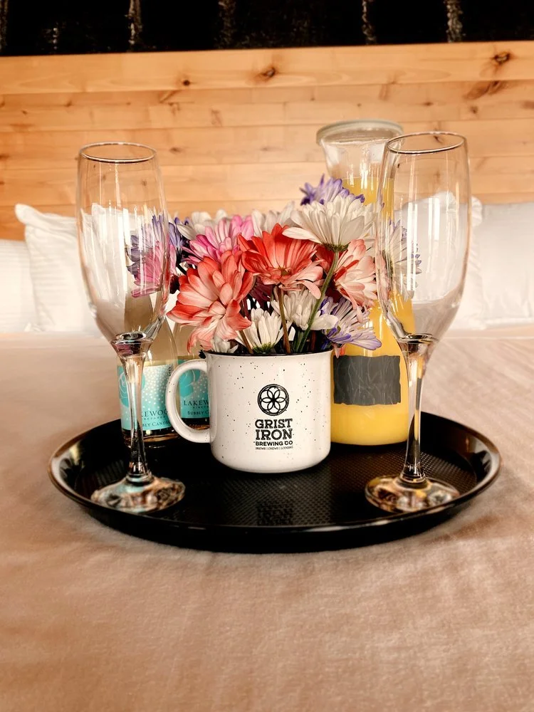 A tray with a mug of flowers, two champagne flutes, a candle, and a pitcher of orange juice on a table with a beige tablecloth.