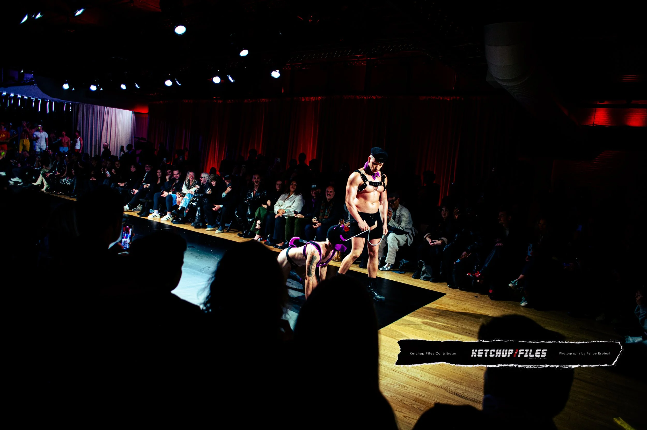 A performer on a runway wearing a leather harness and shorts, holding a leash attached to a person dressed as a dog with purple ears and a collar, during a fashion show or event with an audience watching.