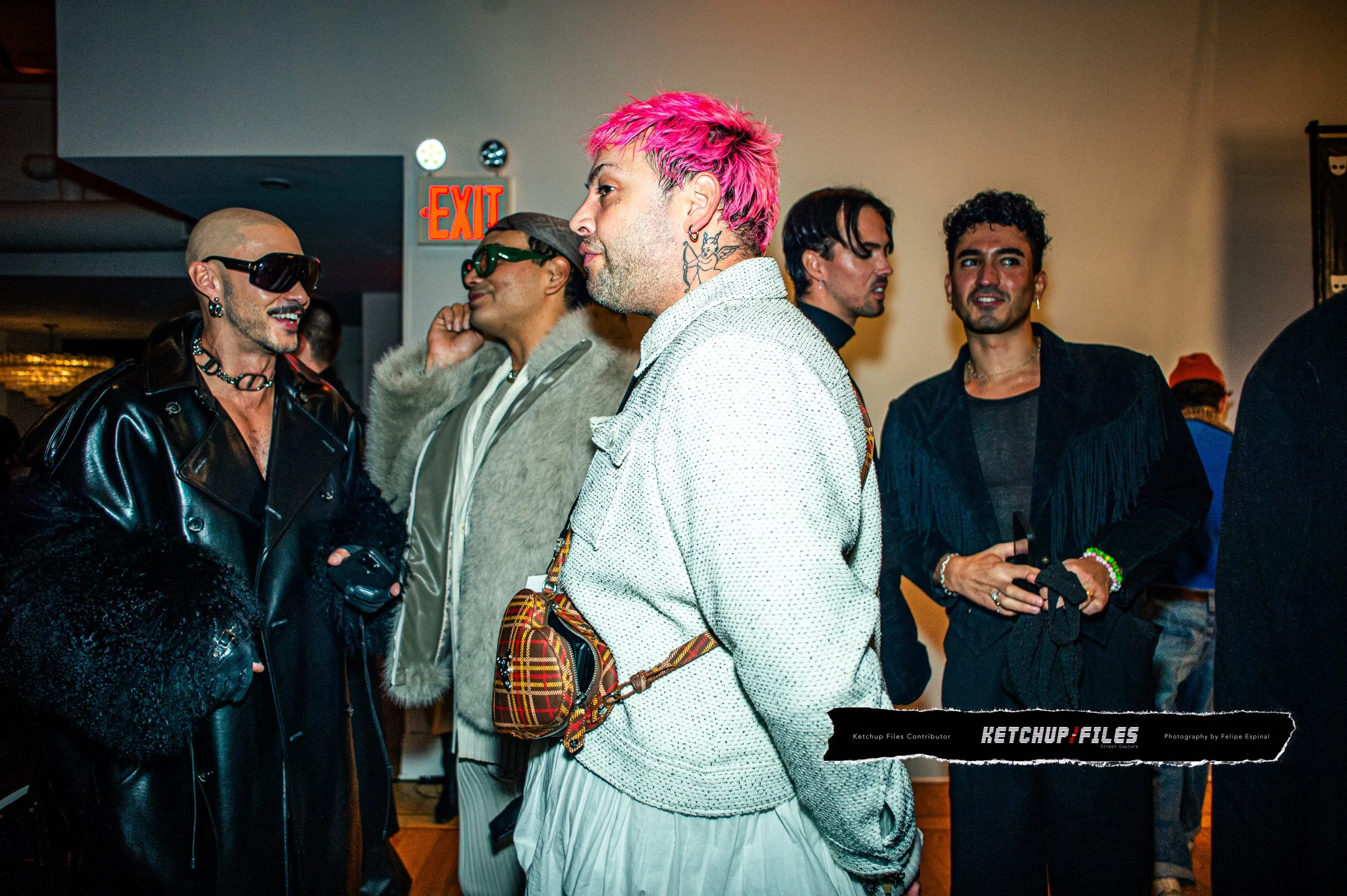 Group of five men with diverse styles socializing indoors, one has pink hair, another in a leather jacket, some wearing sunglasses, with an exit sign in the background.