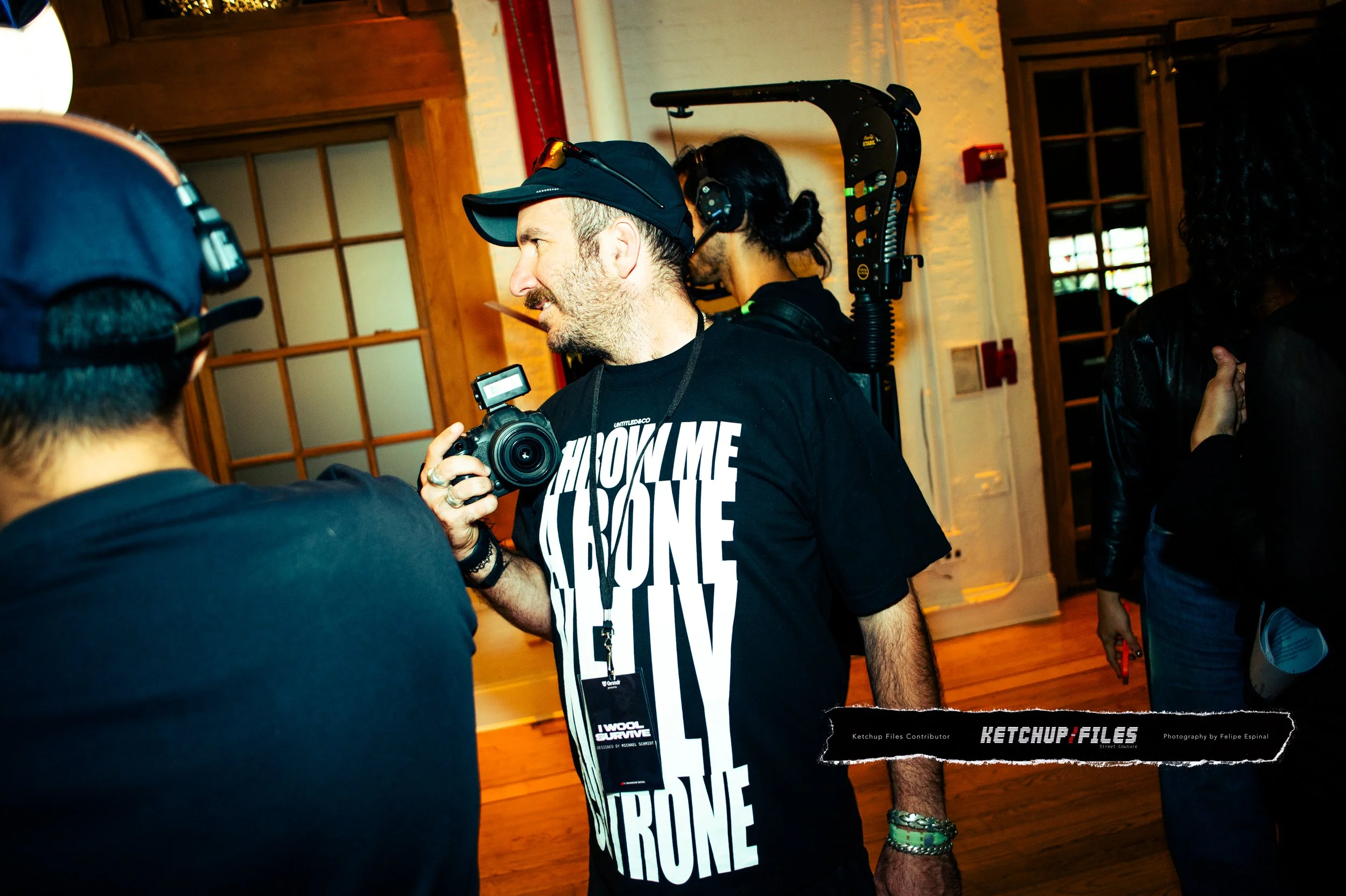 A man wearing a black cap, black t-shirt with white text, and wristbands is holding a camera and talking to others in an indoor setting with wooden floors and large windows.