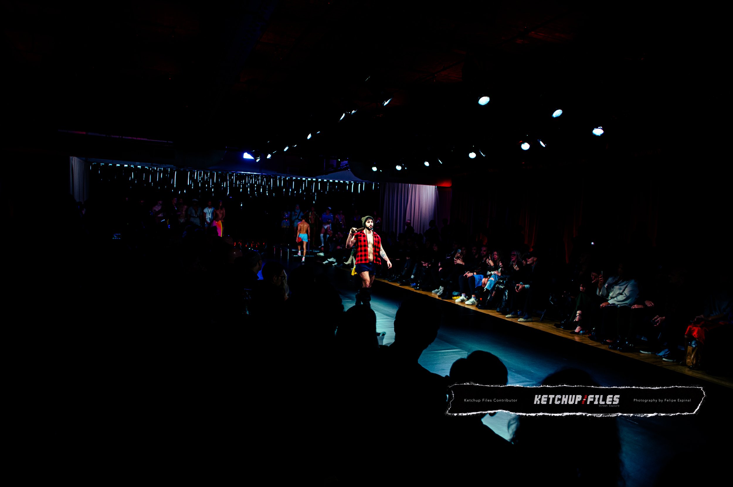 Models walk on a runway during a fashion show, with audience seated on both sides of the runway and stage lighting overhead.