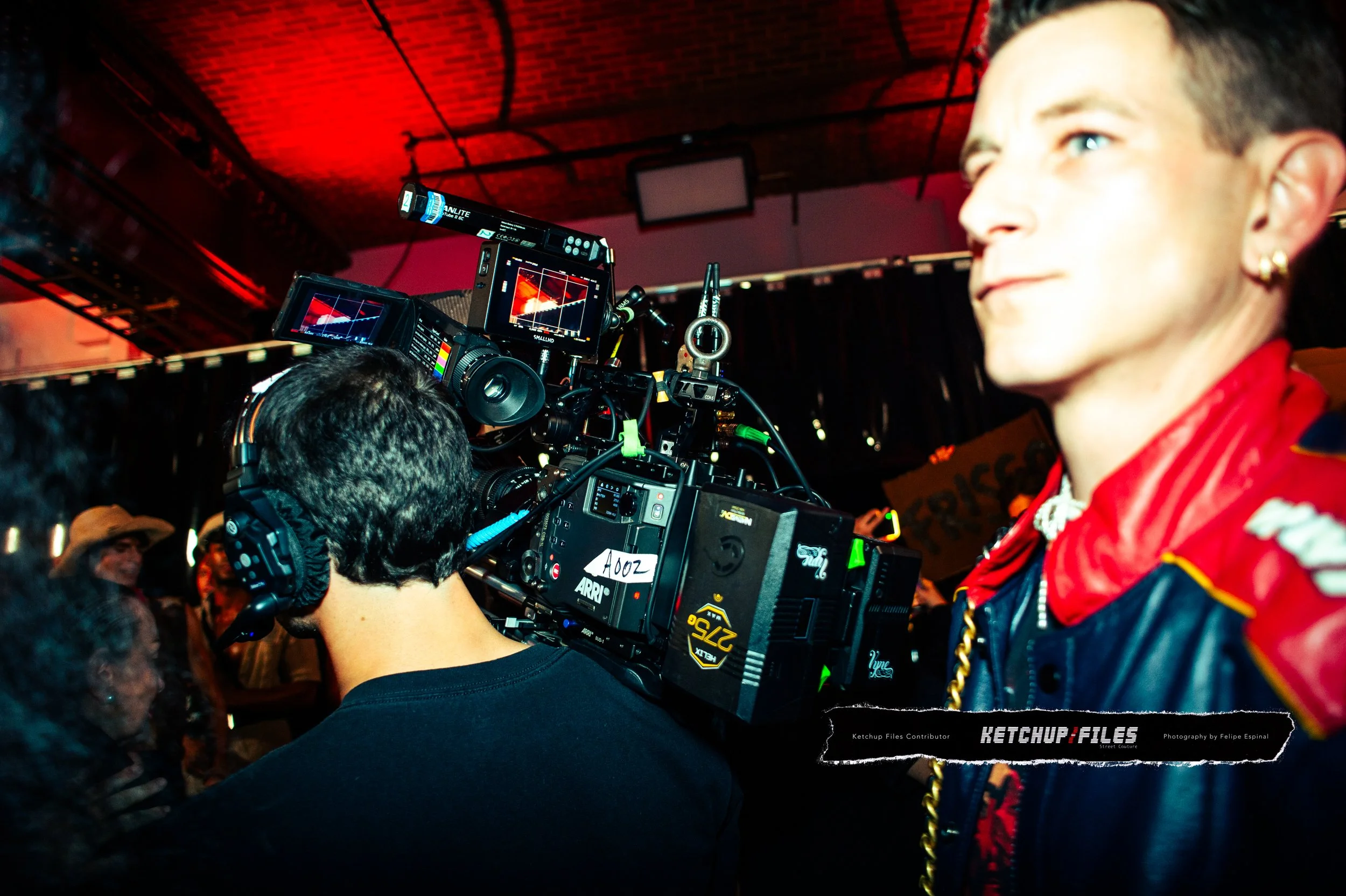 A woman with short dark hair, wearing a red and blue jacket with gold accents, is standing near a professional film camera in a dimly lit venue with a red ceiling. There are people in the background.