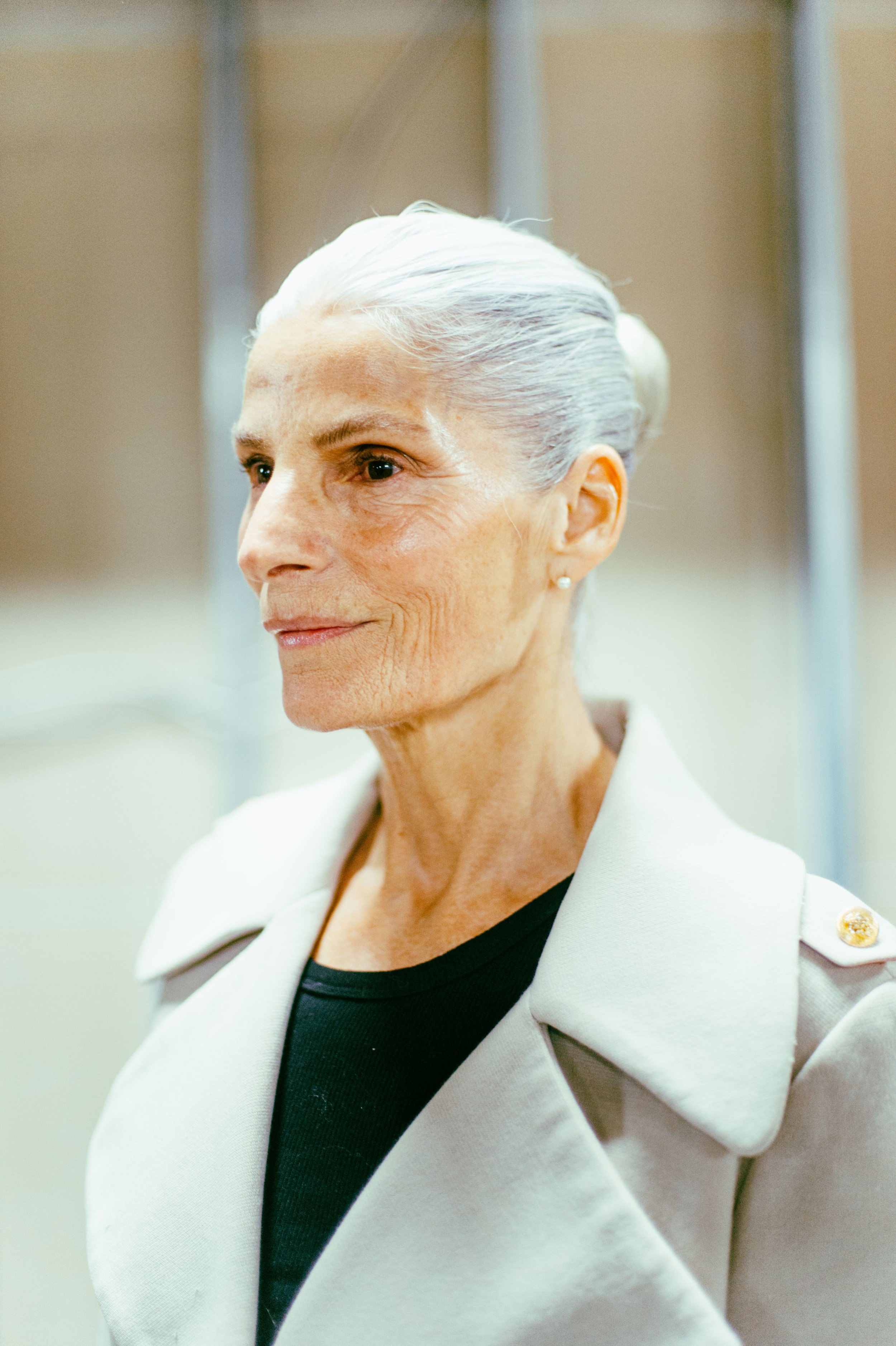 Elderly woman with white hair in a bun, wearing a beige coat, facing sideways.