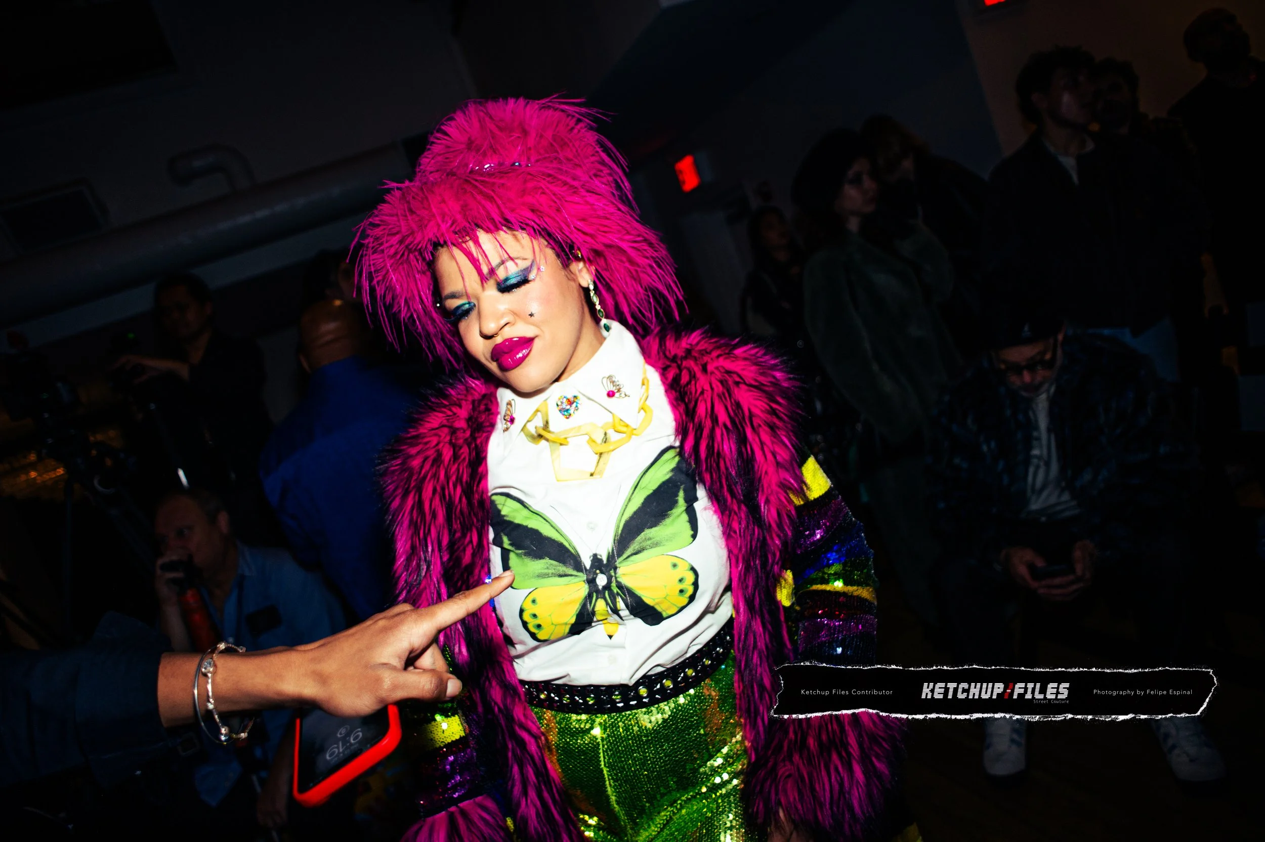 A woman with bright pink hair and makeup, wearing a colorful outfit with a butterfly design on her shirt, a large necklace, and a chunky coat, standing in a crowded indoor space.