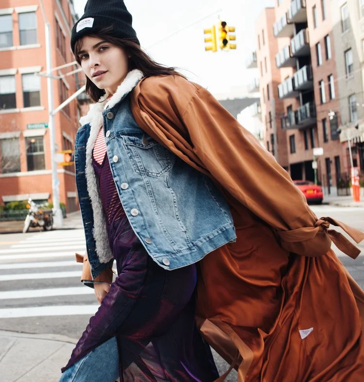 Young woman walking on city street at crosswalk, wearing a black beanie, denim jacket with shear lining, purple dress, and brown long coat, with modern apartment buildings and traffic lights in the background.