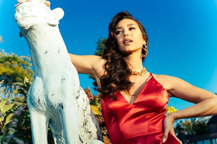 A woman in a red dress with gold jewelry standing outdoors next to a white horse sculpture under a clear blue sky.