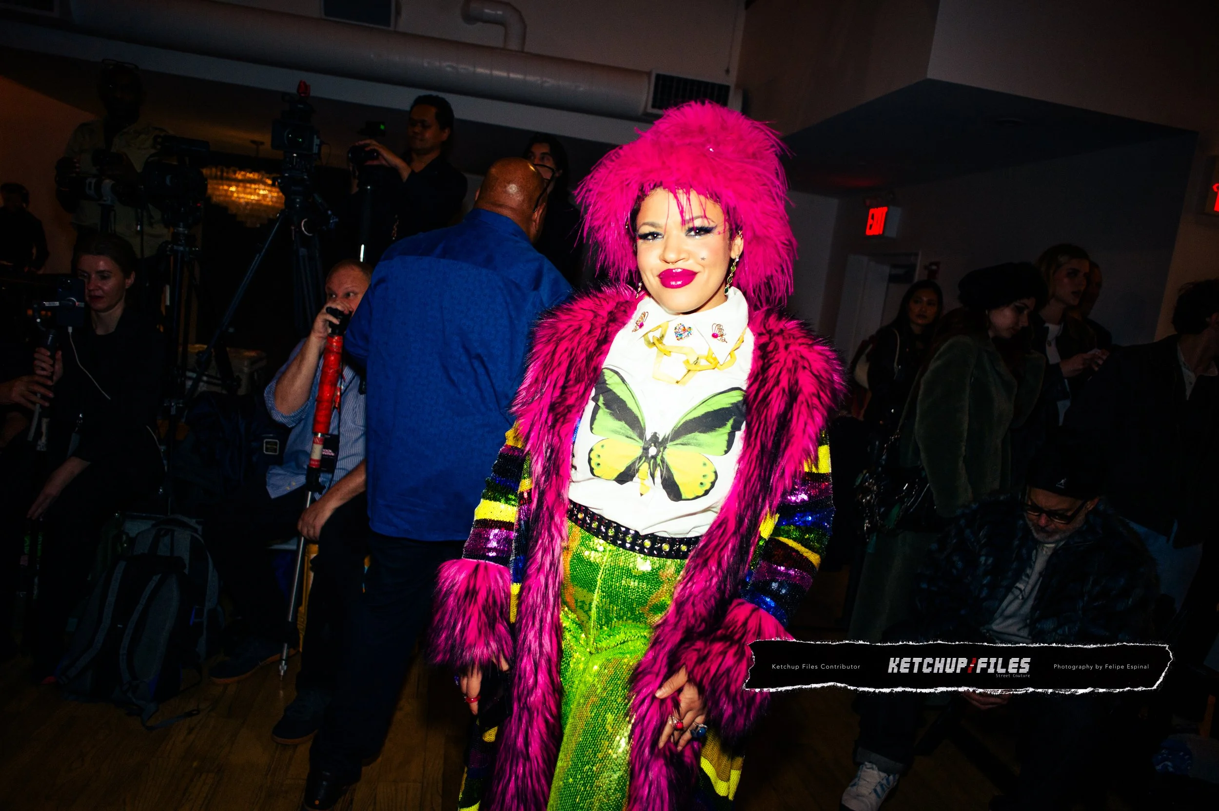A woman with bright pink hair and makeup is smiling at a social gathering, wearing a colorful outfit with a butterfly graphic shirt, striped and sequined sleeves, and a pink furry coat. There are people and cameras in the background.
