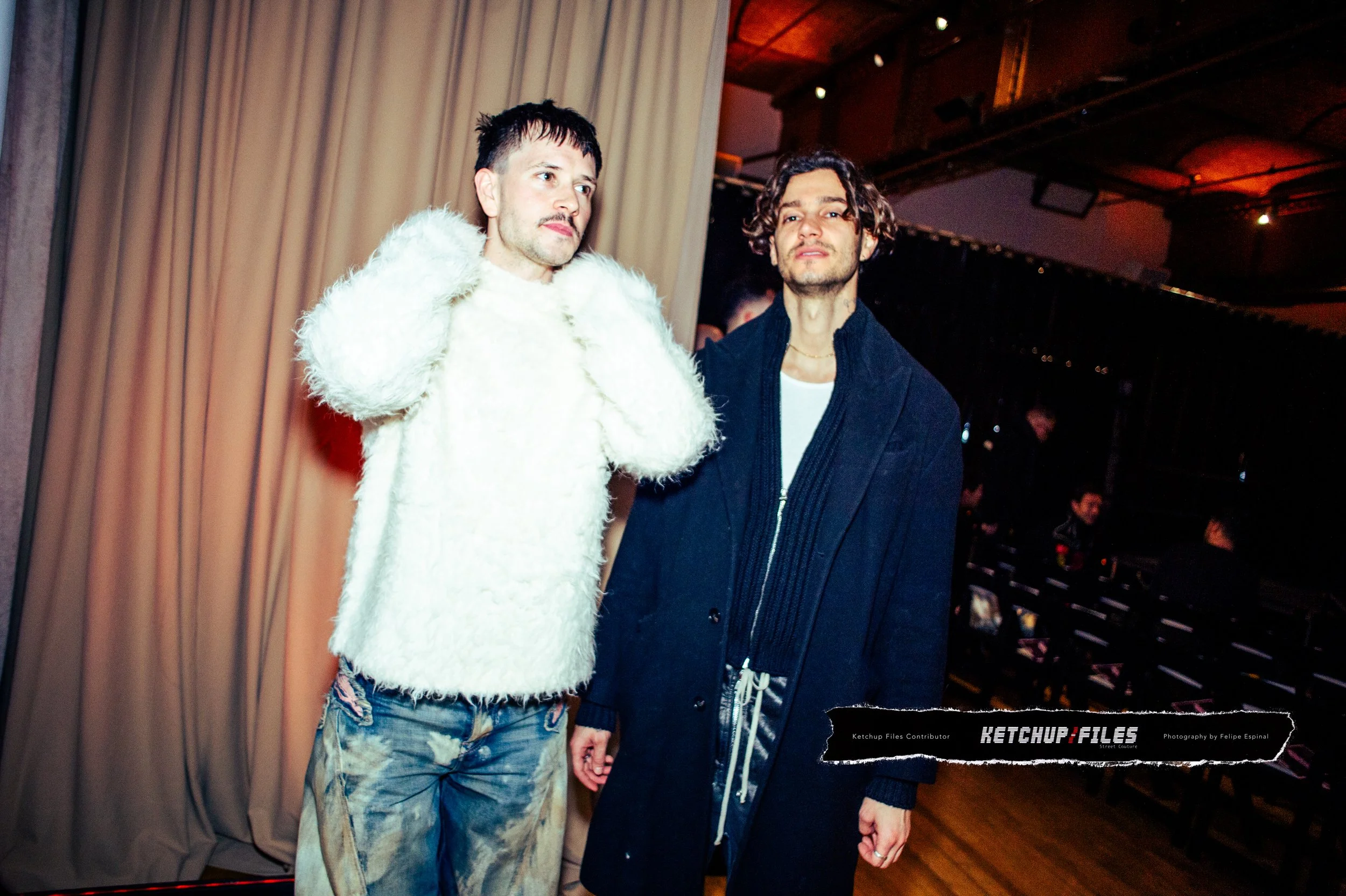 Two men standing side by side indoors, one wearing a white fuzzy sweater and distressed jeans, the other in a dark coat and hoodie, with chairs and a curtain in the background.