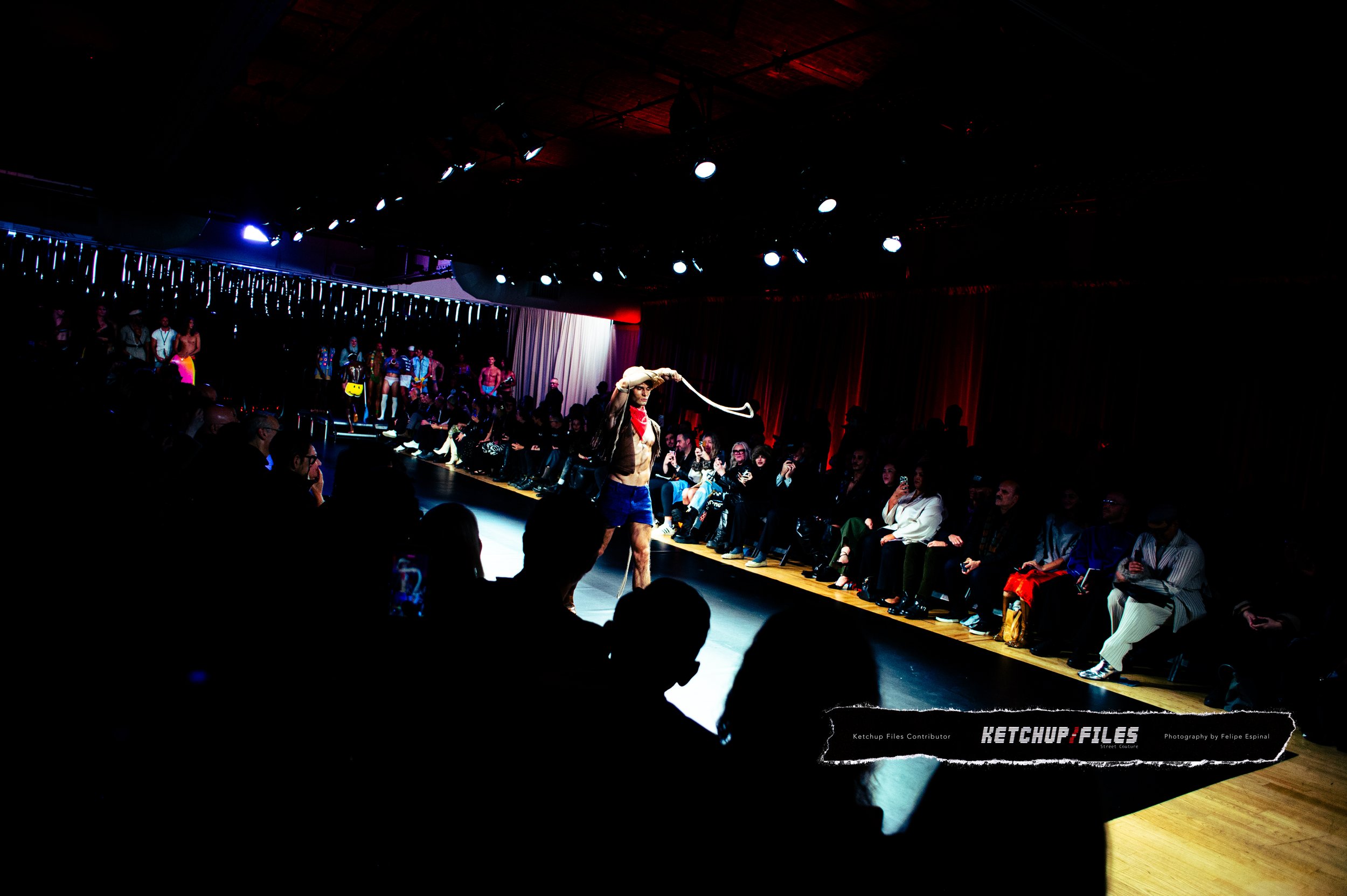 A fashion runway show with a male model walking down the runway as an audience watches from both sides. The setting is a dark room with spotlights and decorative hanging lights. The model is dressed in shorts and a jacket, holding a whip, and is posi