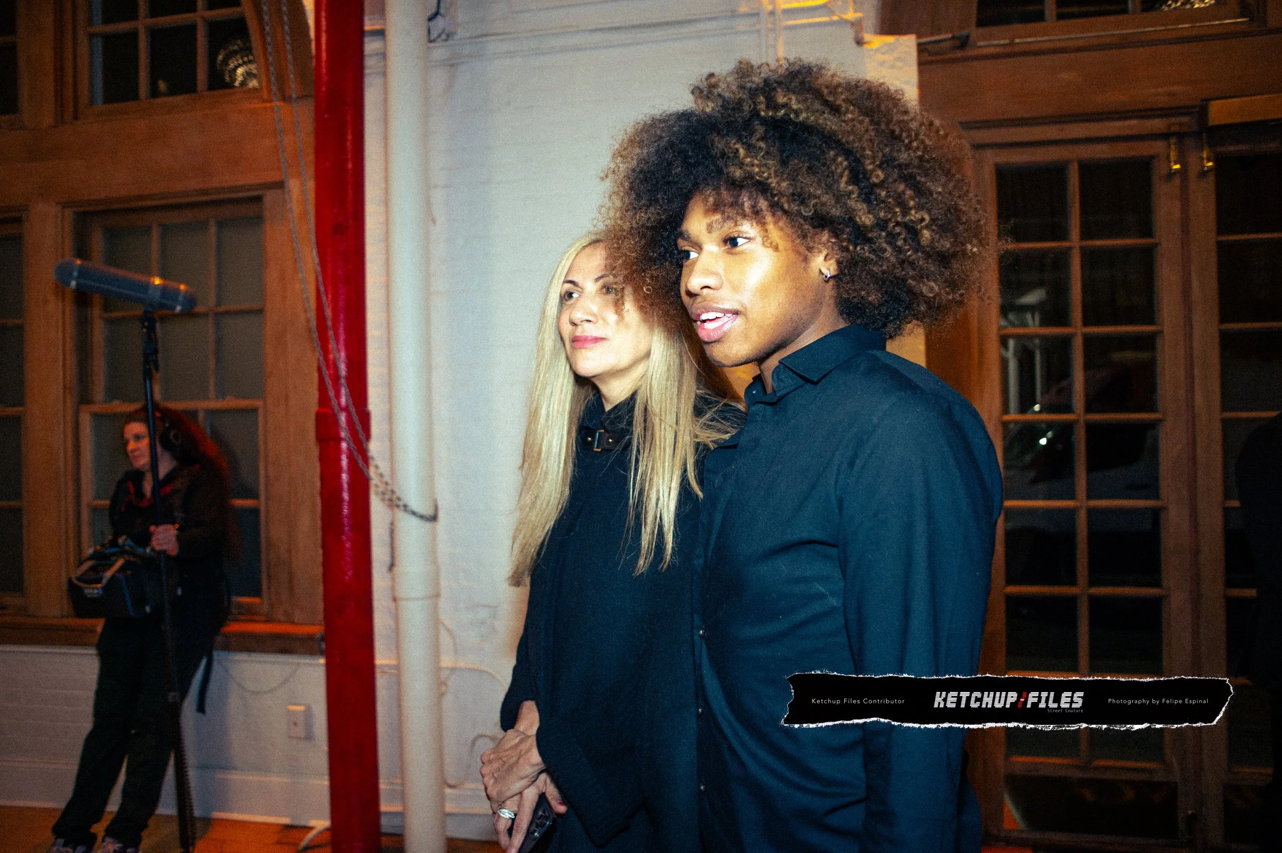 Two women standing indoors near a window, with a person operating a film camera on a tripod in the background.