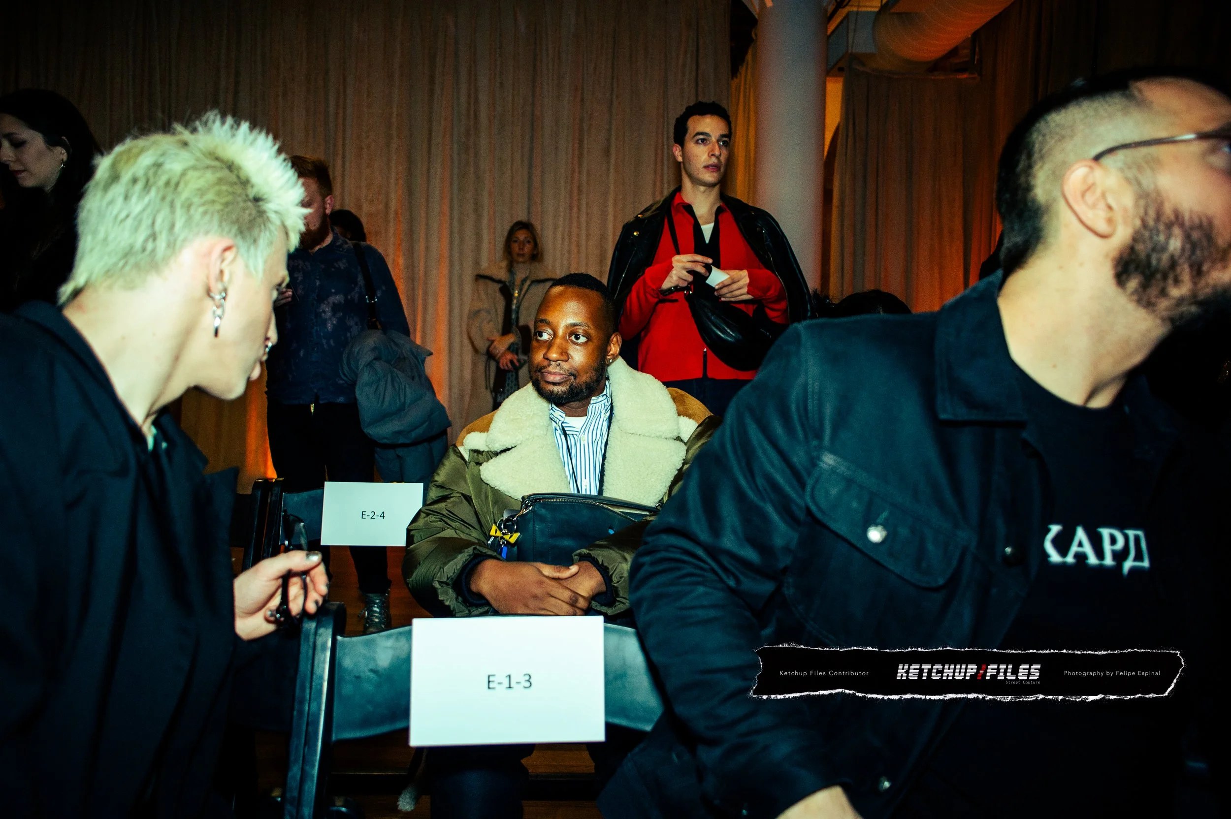 A group of people seated at a conference, with some looking at each other and others standing in the background, including a man with a green fur-lined jacket sitting at a table labeled E-1-3 and a woman with bleached blonde hair on the left.