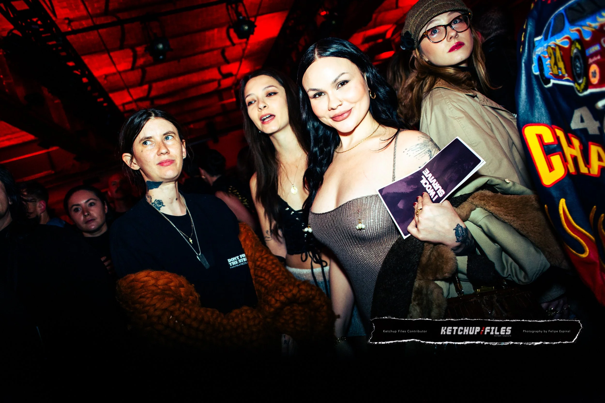 Group of women at a dark indoor event with red lighting, some looking at the camera, one holding a flyer, in casual and stylish clothing.