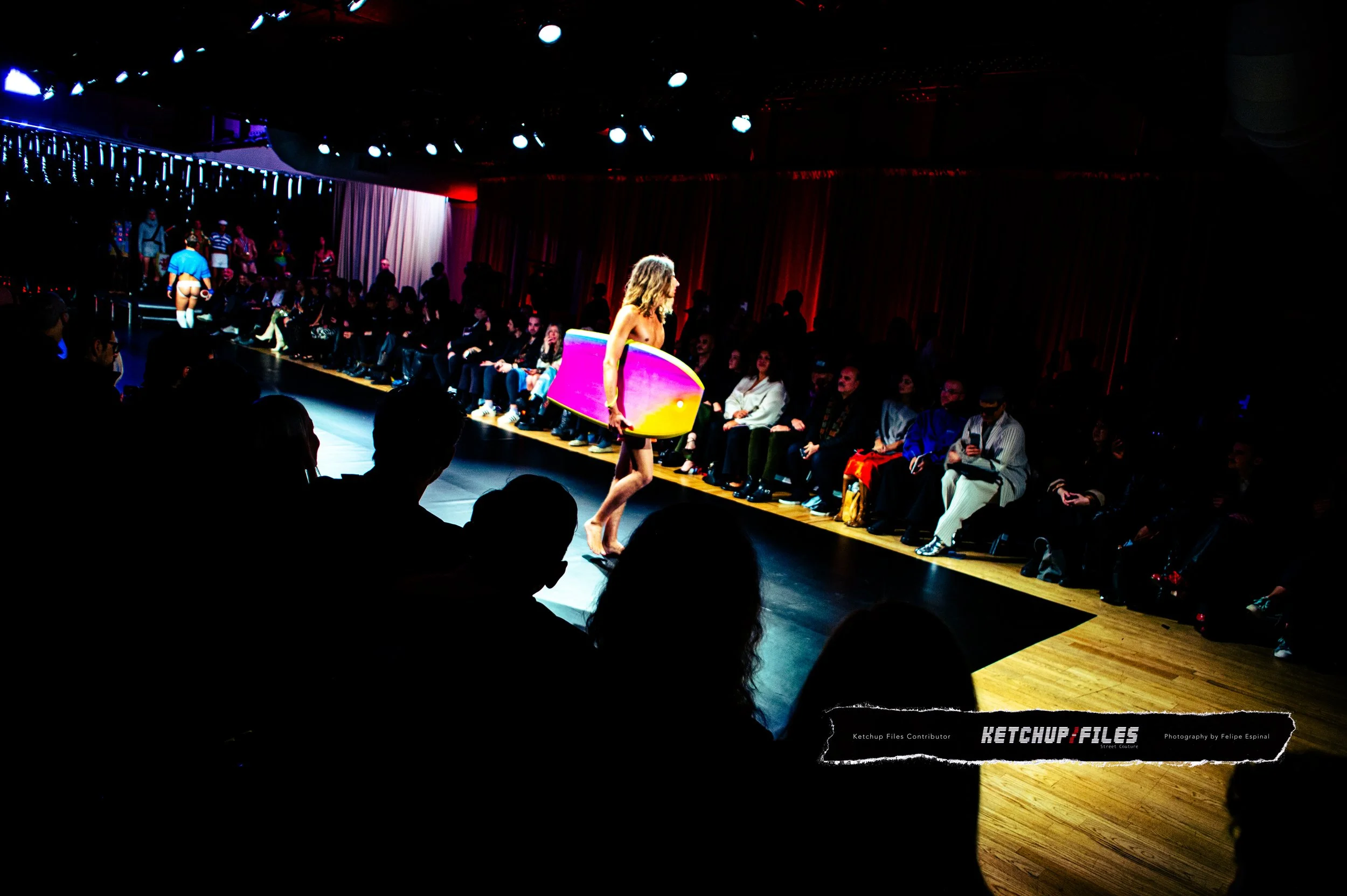 A woman walking on a runway at a fashion show, carrying a pink, purple, and yellow surfboard. The audience is seated along the sides, watching the show in a dimly lit room with ceiling lights and red curtains.