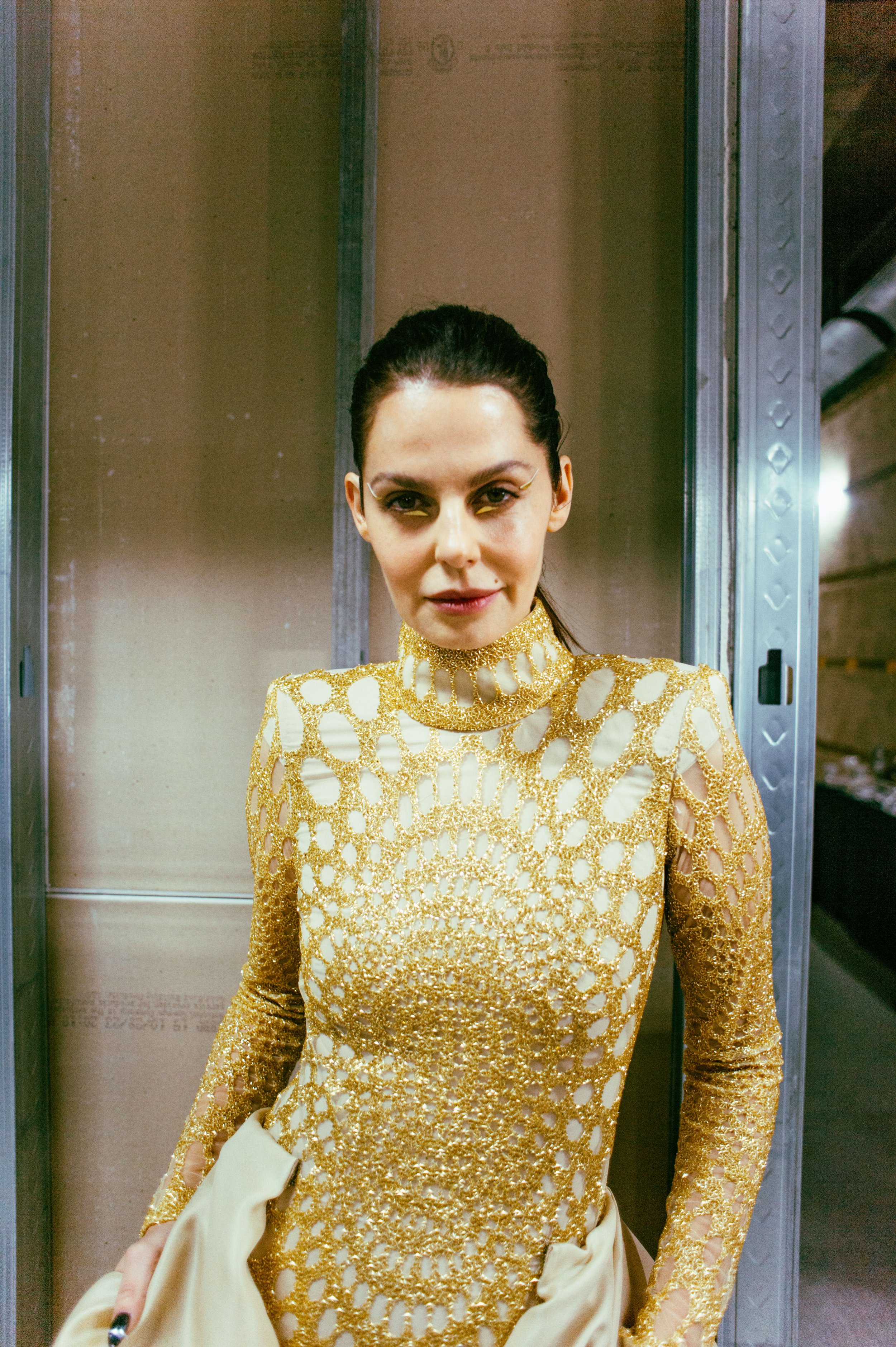 Woman in gold lace dress with intricate pattern, low-lit background.