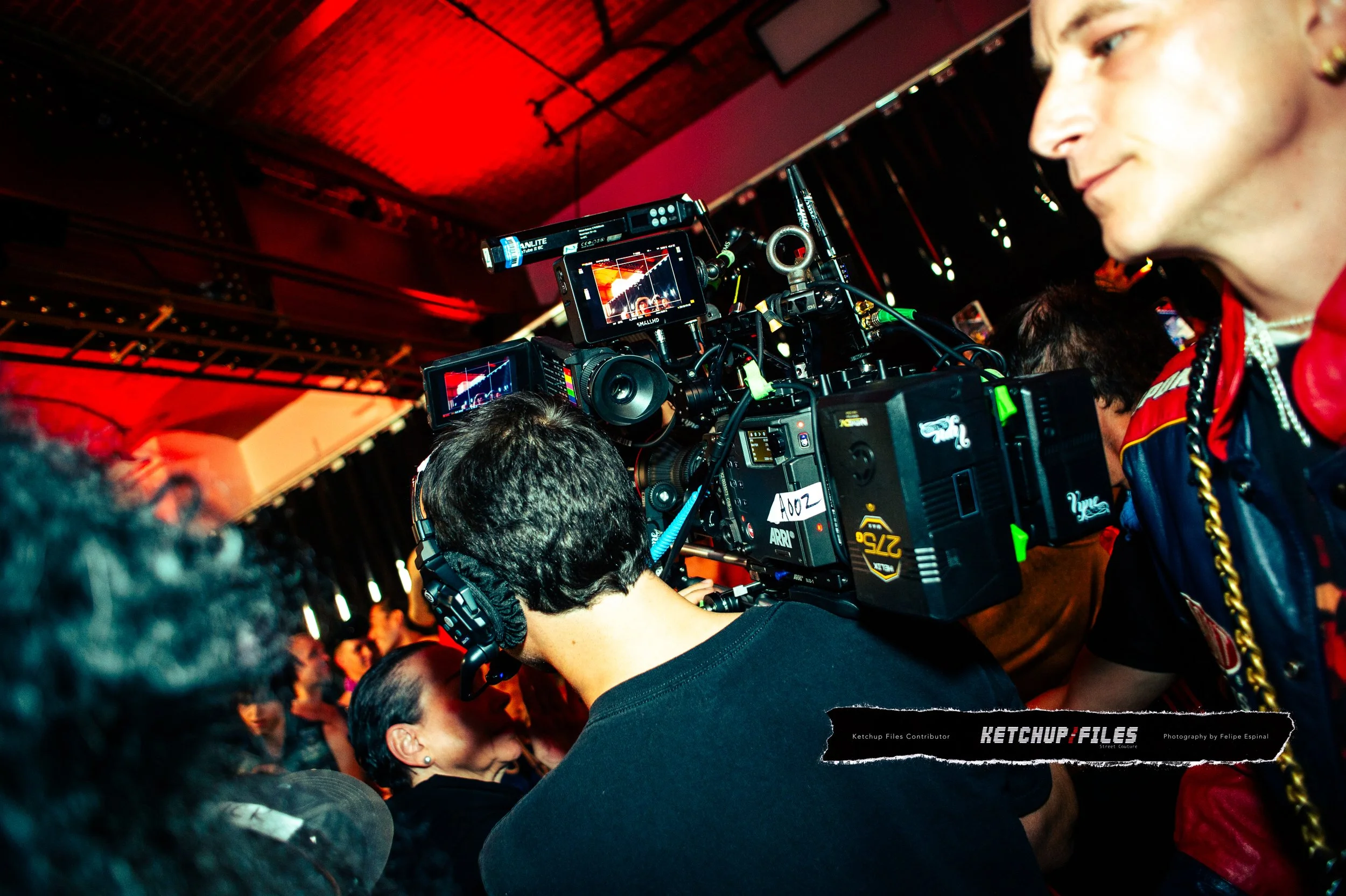 A person operating a professional video camera at an indoor event with a crowd in the background and red stage lighting.