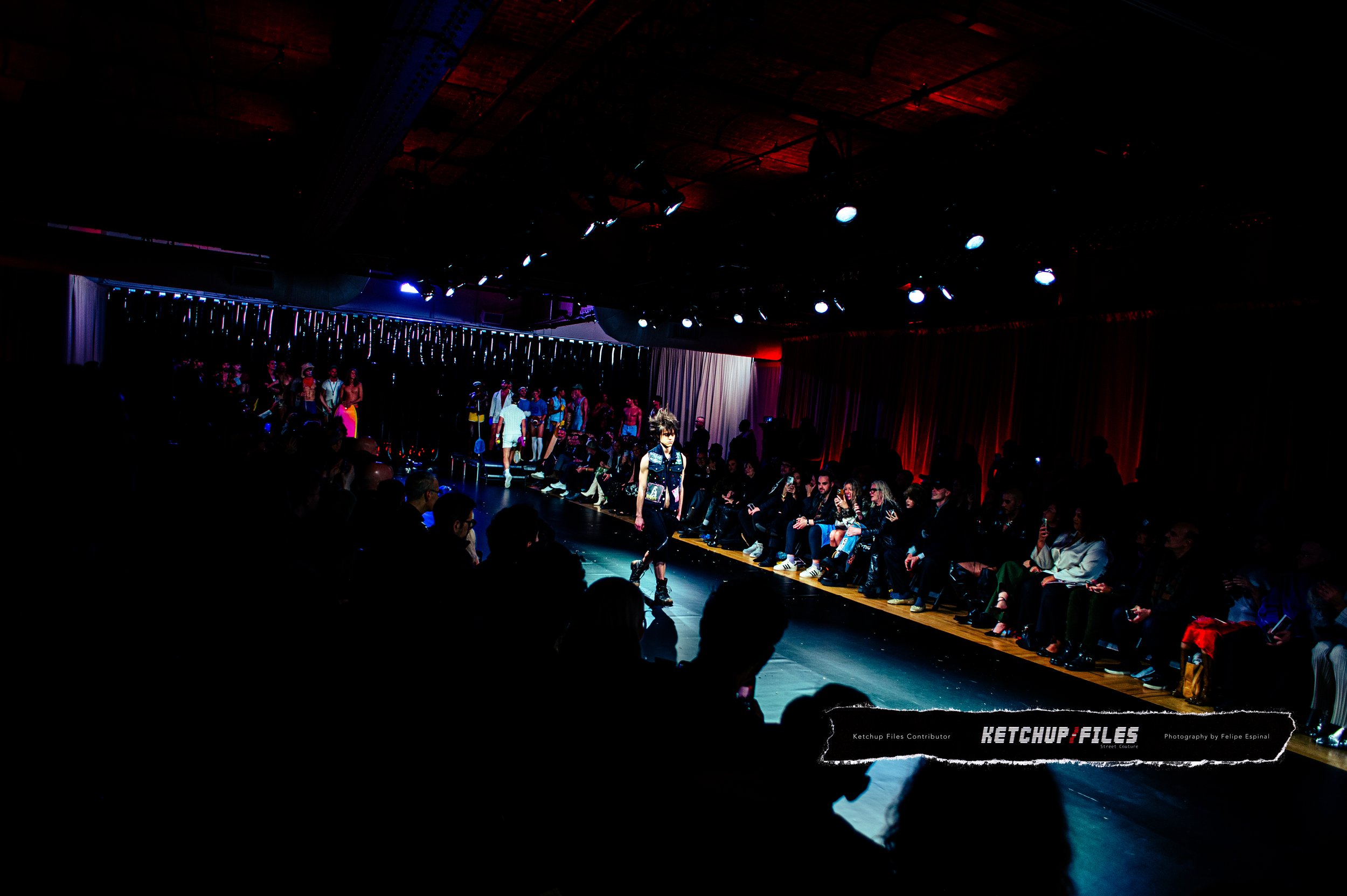 Fashion show runway with models walking and audience seated on both sides, dim lighting with colorful stage lights.