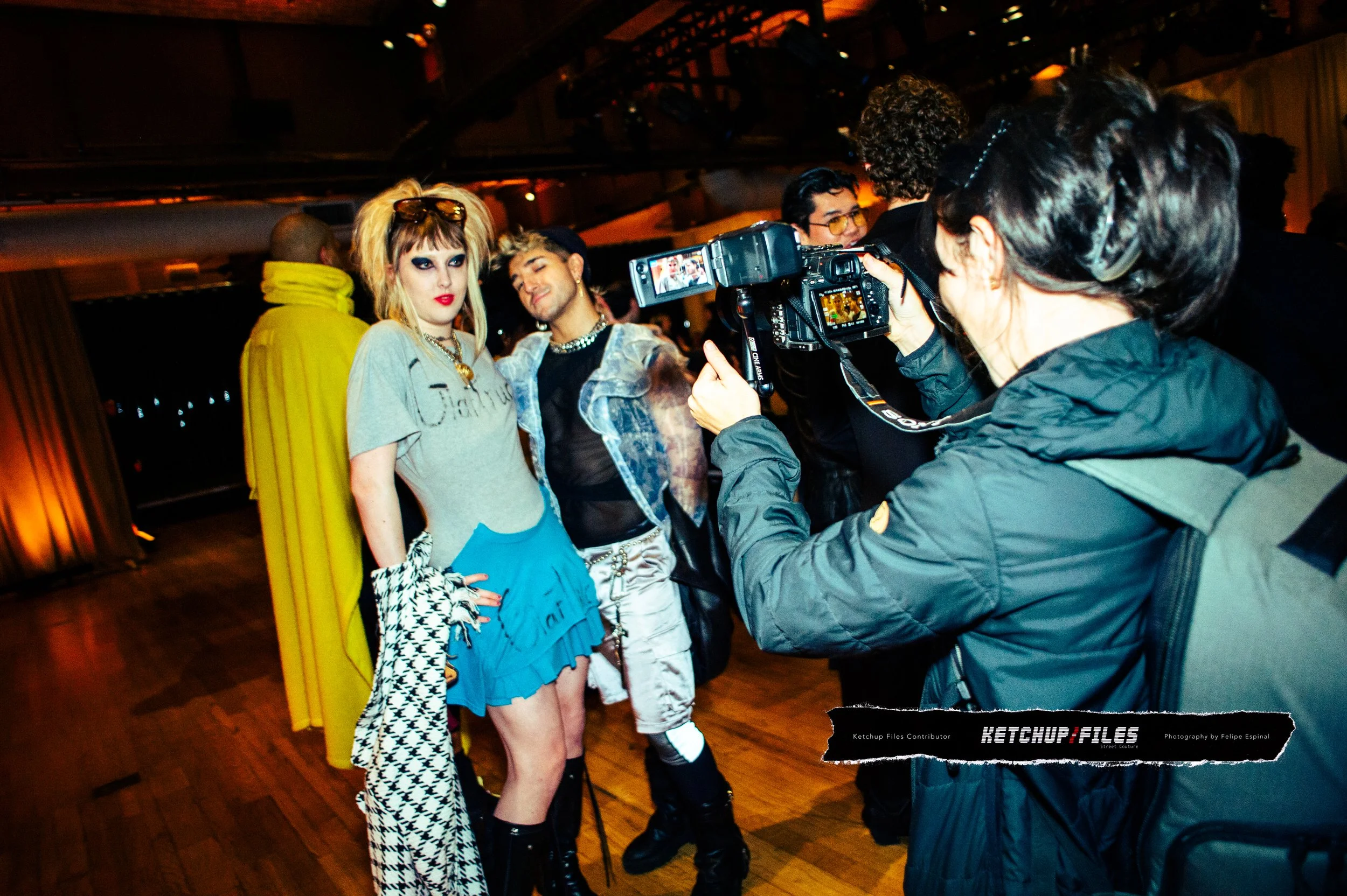 People posing for a photo at an indoor event, with one person taking a picture. The group includes a woman with blonde hair and makeup wearing a Gucci t-shirt, and another person with an edgy style. The background is dimly lit with warm lighting.