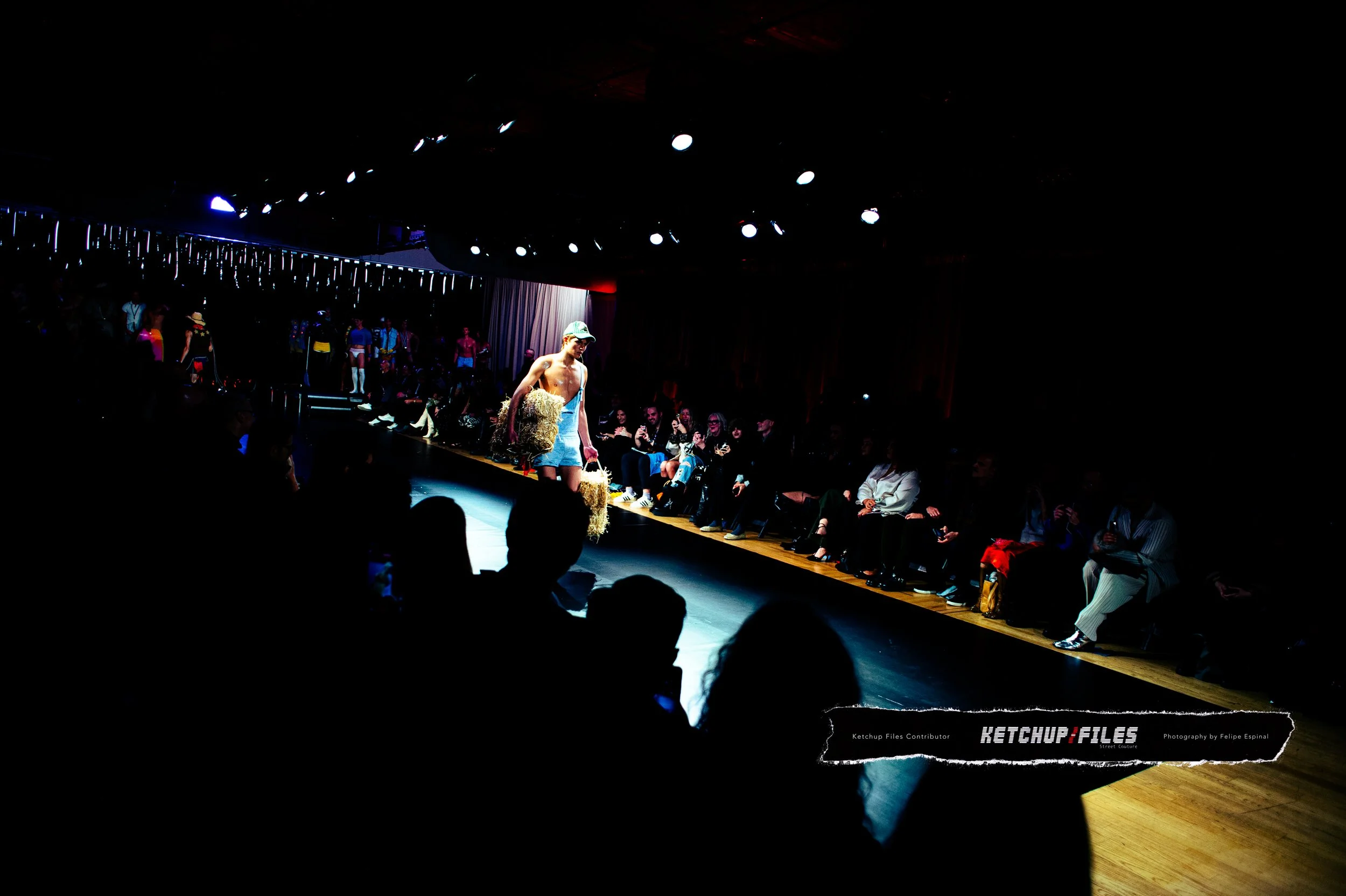 A model walking down a runway at a fashion show, holding a straw hat, with an audience seated on either side in a dark, indoor venue with stage lights overhead.