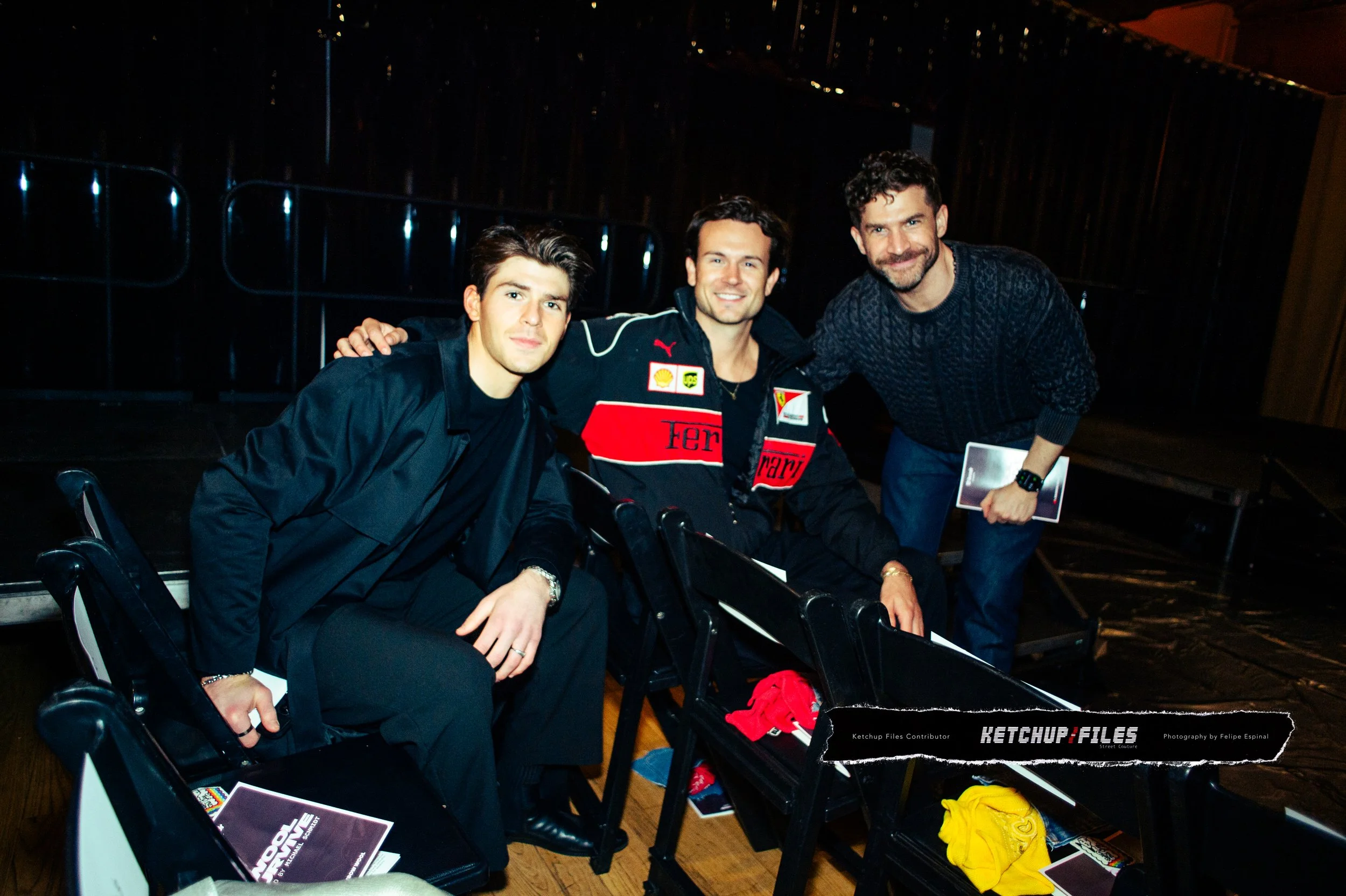 Three men sitting and standing together in a dimly lit room, one wearing a Ferrari racing jacket, two dressed in casual dark clothing, smiling for the camera.