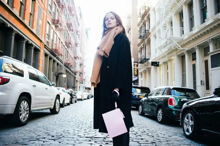 A woman standing on a cobblestone street in an urban area with parked cars and buildings on both sides, wearing a black coat and a tan scarf, holding a pink shopping bag.