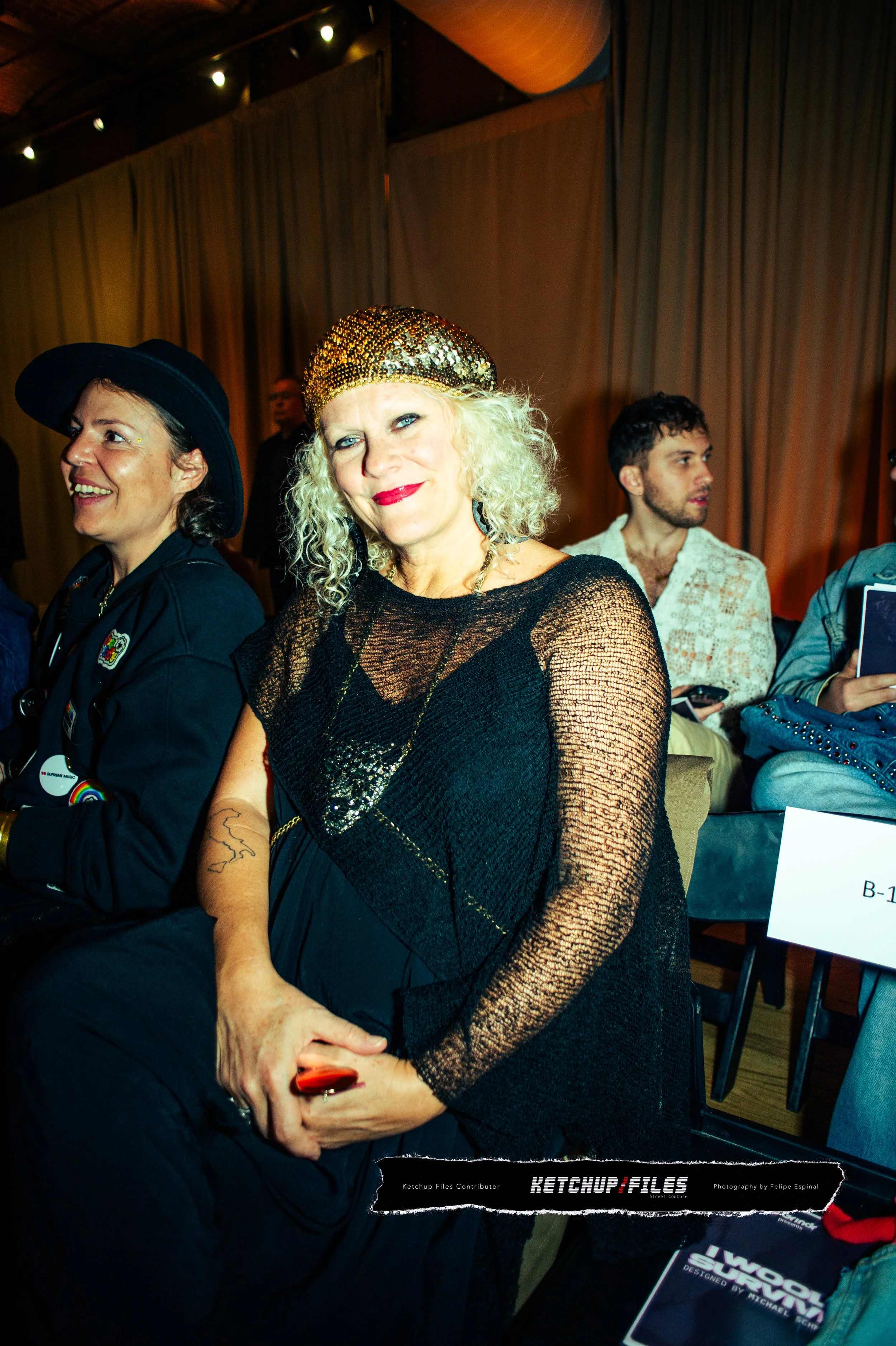 A woman with curly blonde hair wearing a gold beaded headpiece and a black sheer top, sitting in a crowded event space with beige curtains and other attendees visible in the background.