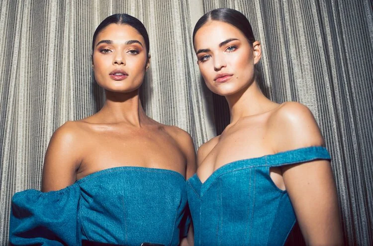 Two women with dark hair and makeup wearing off-the-shoulder blue denim dresses, standing close together in front of a patterned curtain.