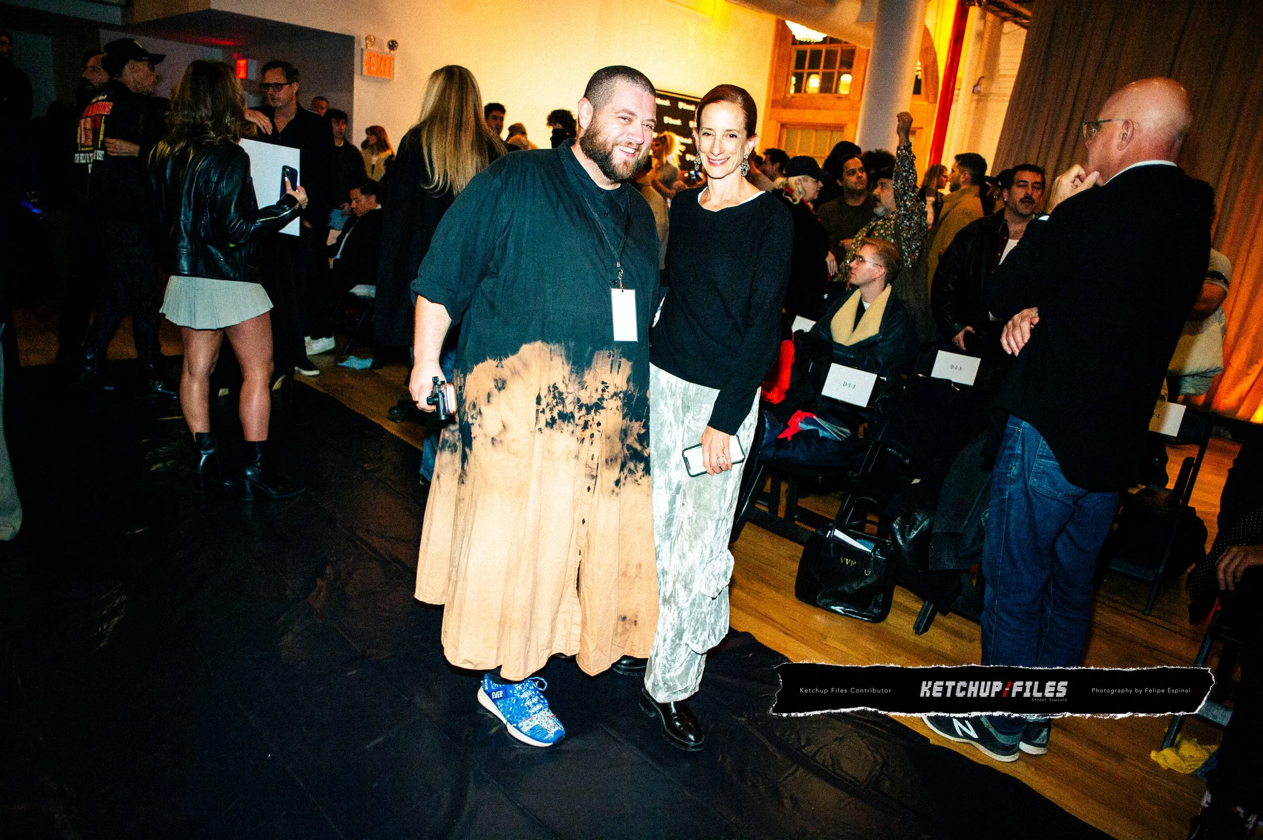 Two people smiling and posing at a crowded indoor event, with many attendees in the background, some seated and some standing, and a woman taking photos.