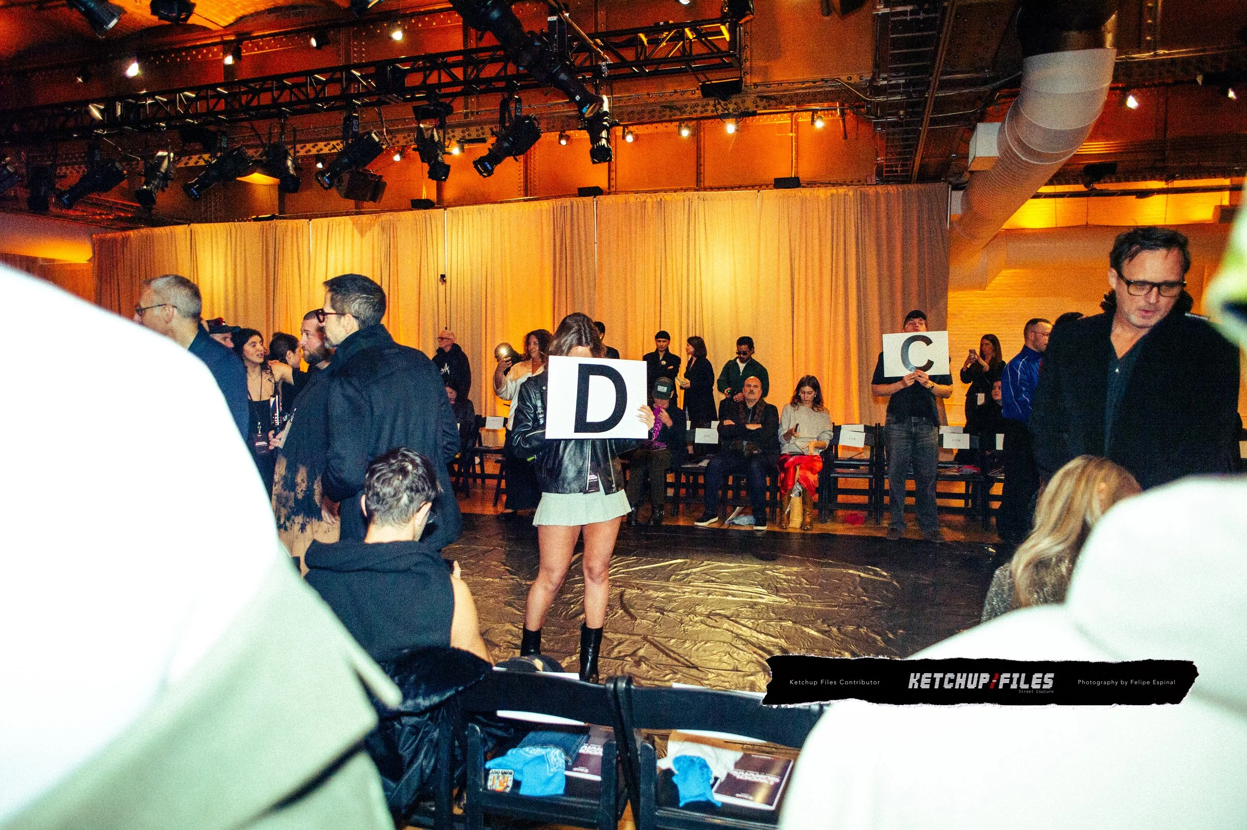 People holding large alphabet signs D and C at an indoor event with a gold curtain backdrop and stage lighting.