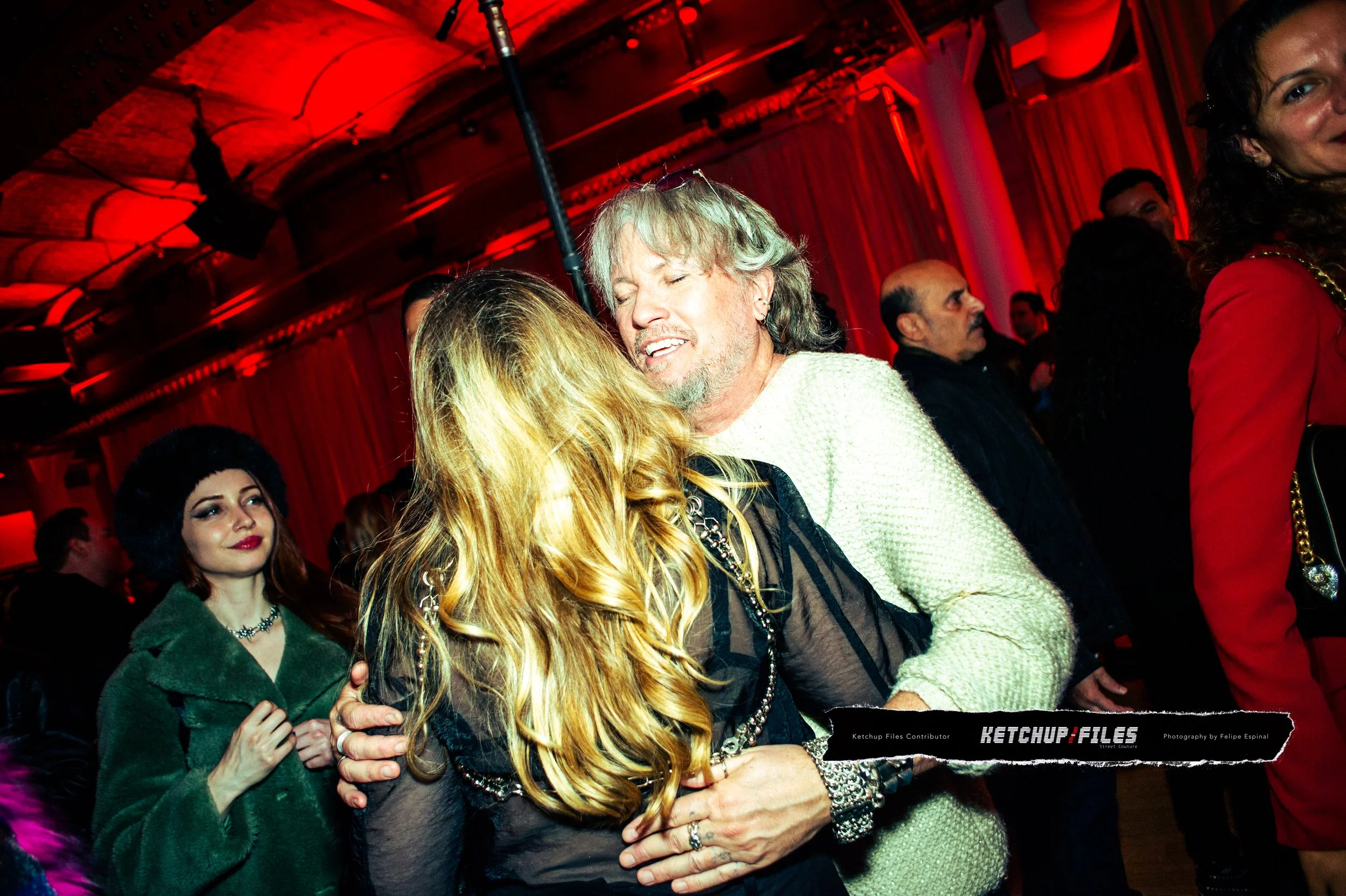 People dancing and socializing at a party in a dimly lit venue with red lighting, including a woman with long blonde hair and a man with gray hair engaged in a close dance.