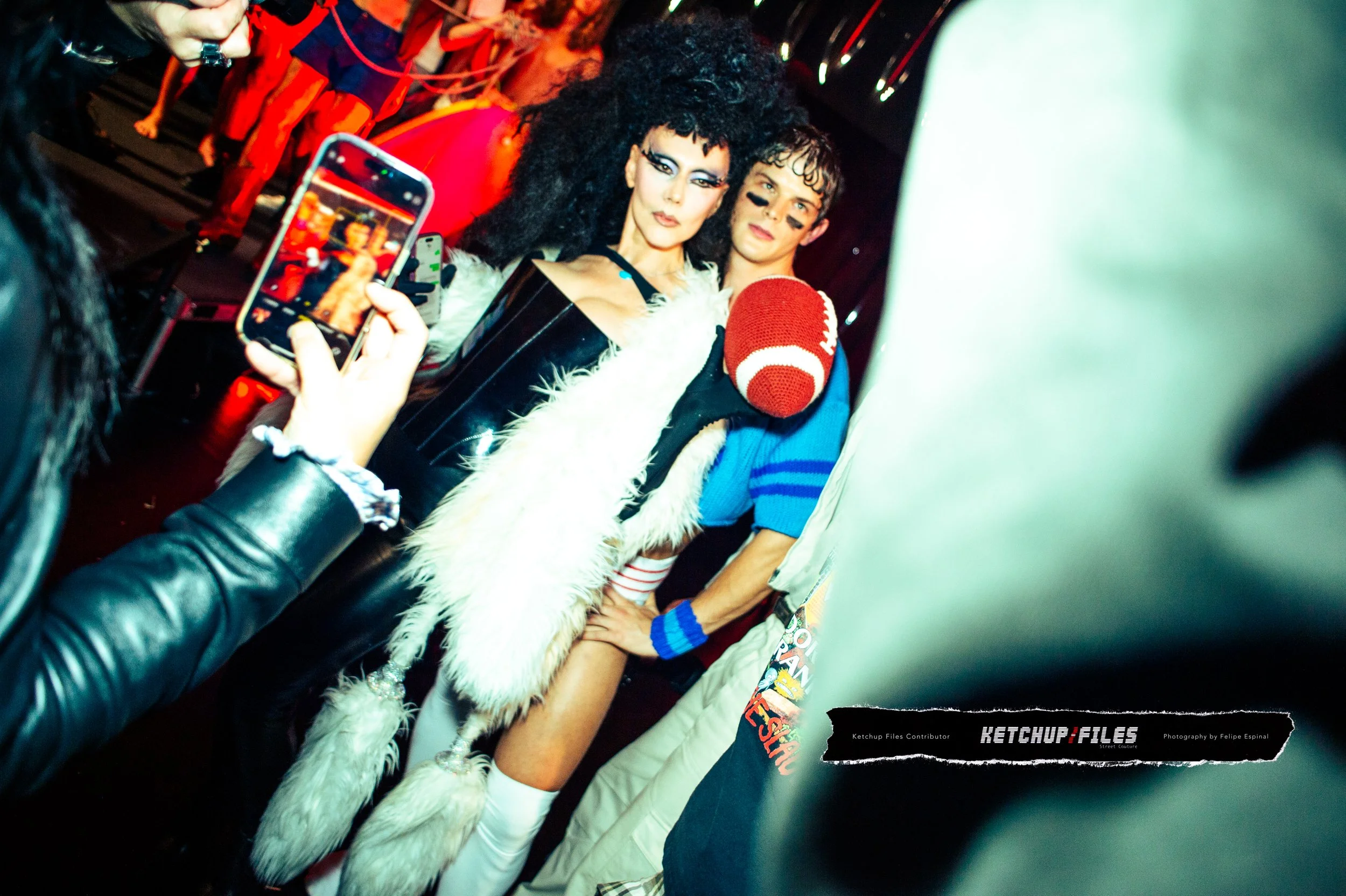Two individuals in costume posing for a photo at a lively event, with one holding a football-shaped prop and another dressed in bold, fashion-forward attire with dark makeup and curly hair. A person in the foreground is taking a picture with a smartp
