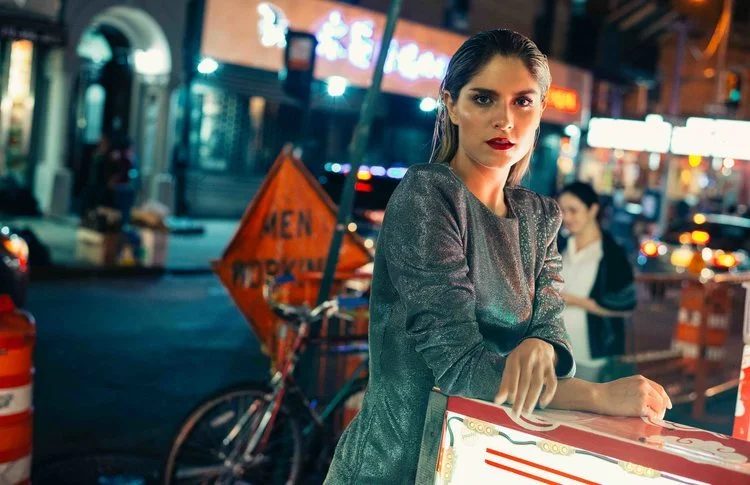 A woman with dark hair, wearing a shiny gray top, stands at a street food cart at night in an urban area with neon signs and pedestrians in the background.