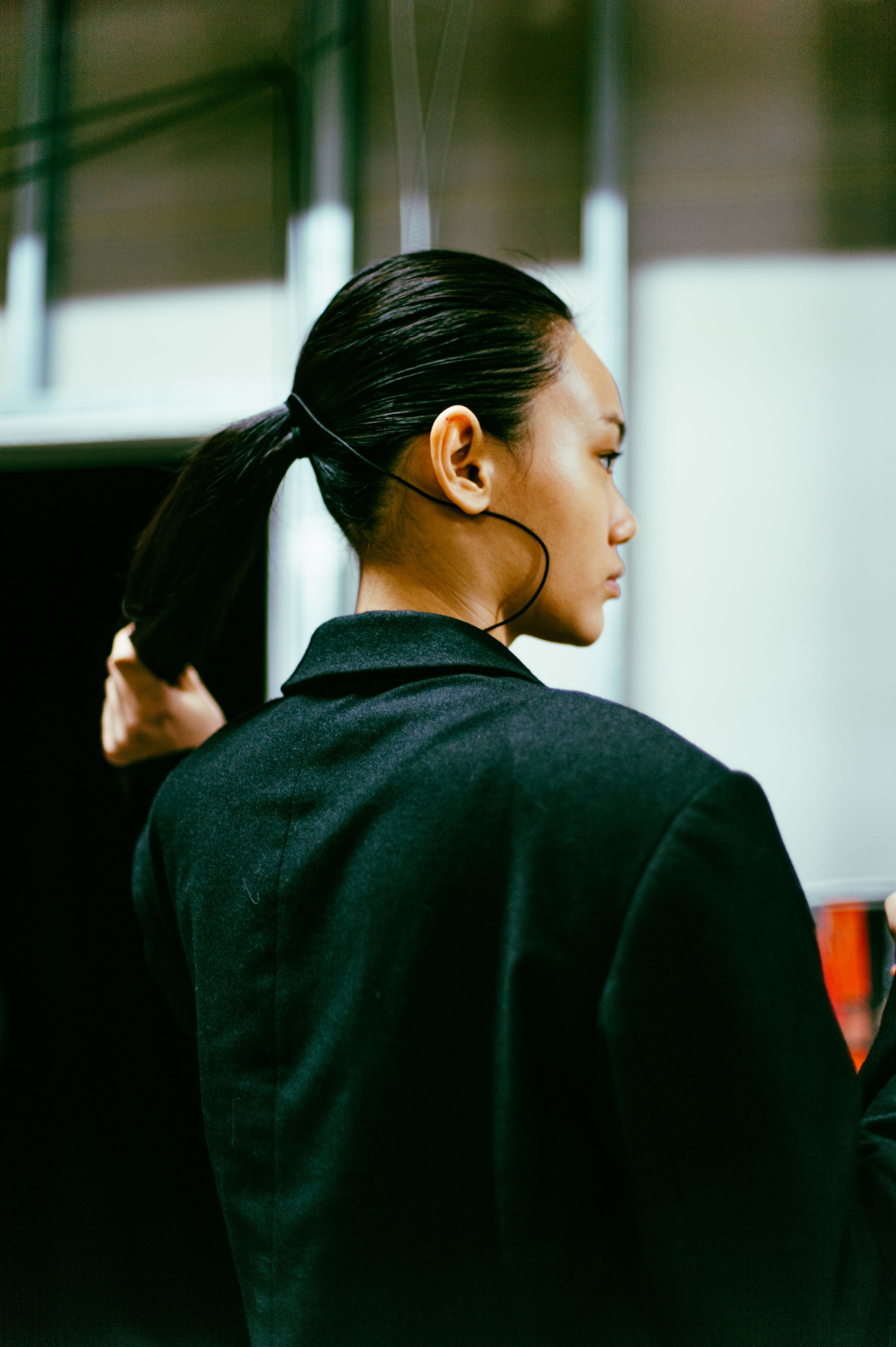Person in a dark blazer with long hair in a ponytail, seen from the back and side, in an indoor setting.