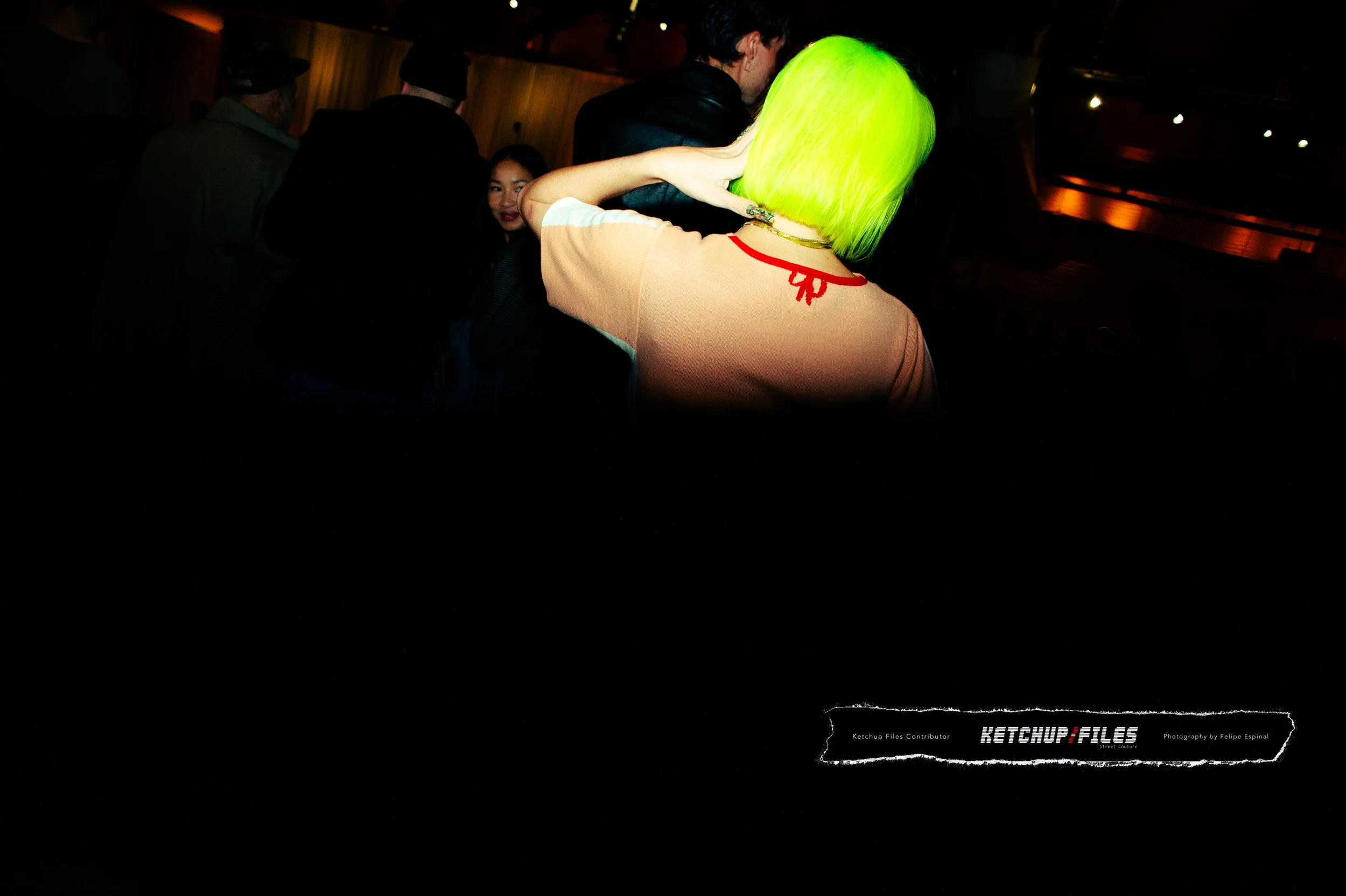 A woman with bright neon green hair taking a photo in a dimly lit indoor setting, with other people in the background.