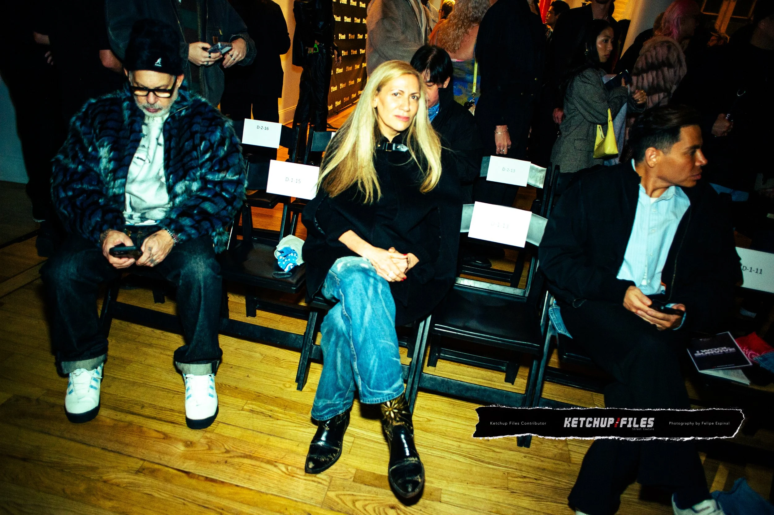 Three individuals seated in a row at a crowded indoor event, with the woman in the middle wearing a black jacket, blue jeans, and shiny black boots, and the two men on either side of her using smartphones.