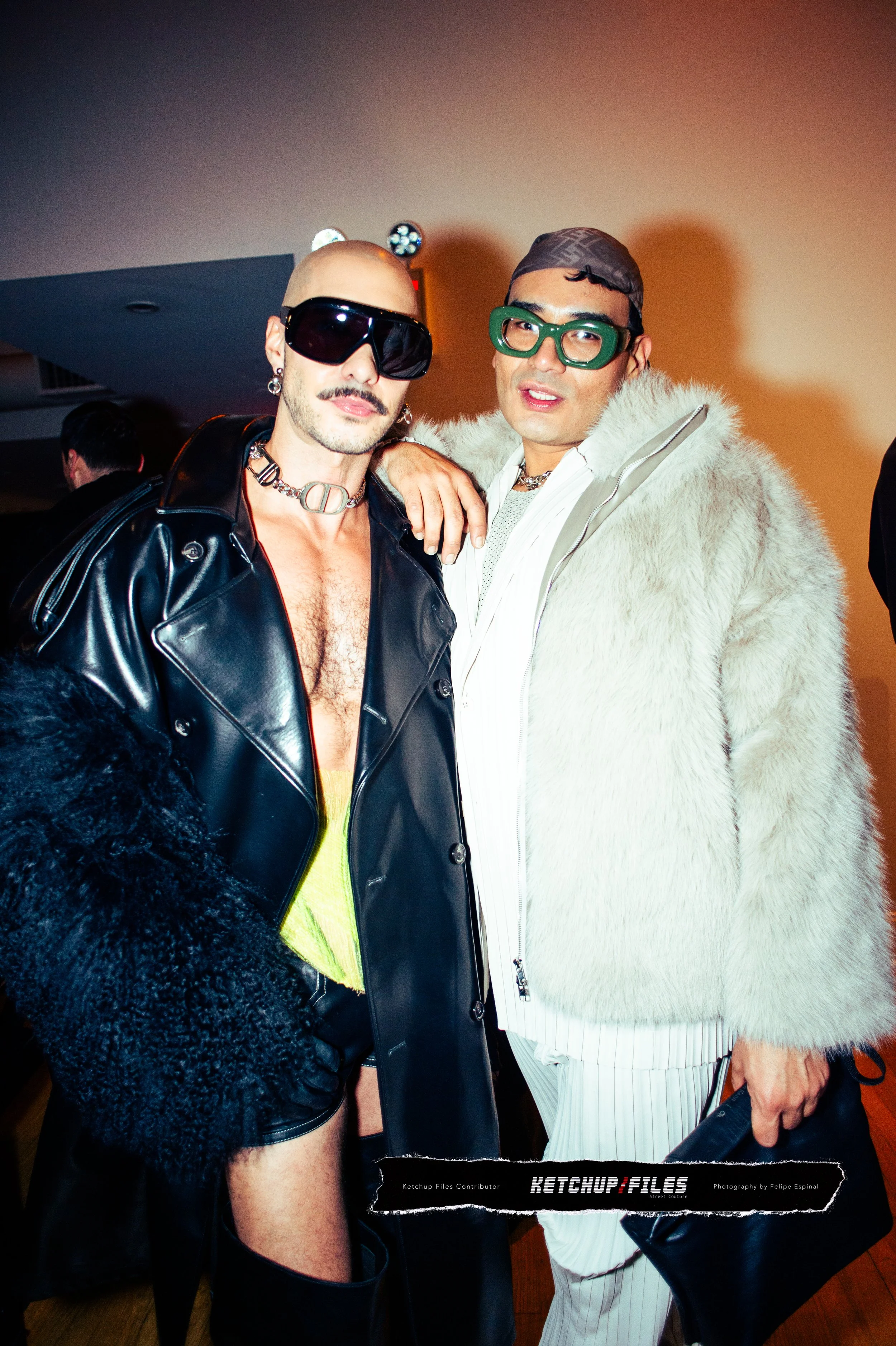 Two individuals dressed in bold, fashionable outfits at a social event, both wearing sunglasses and standing close together in front of a plain background.