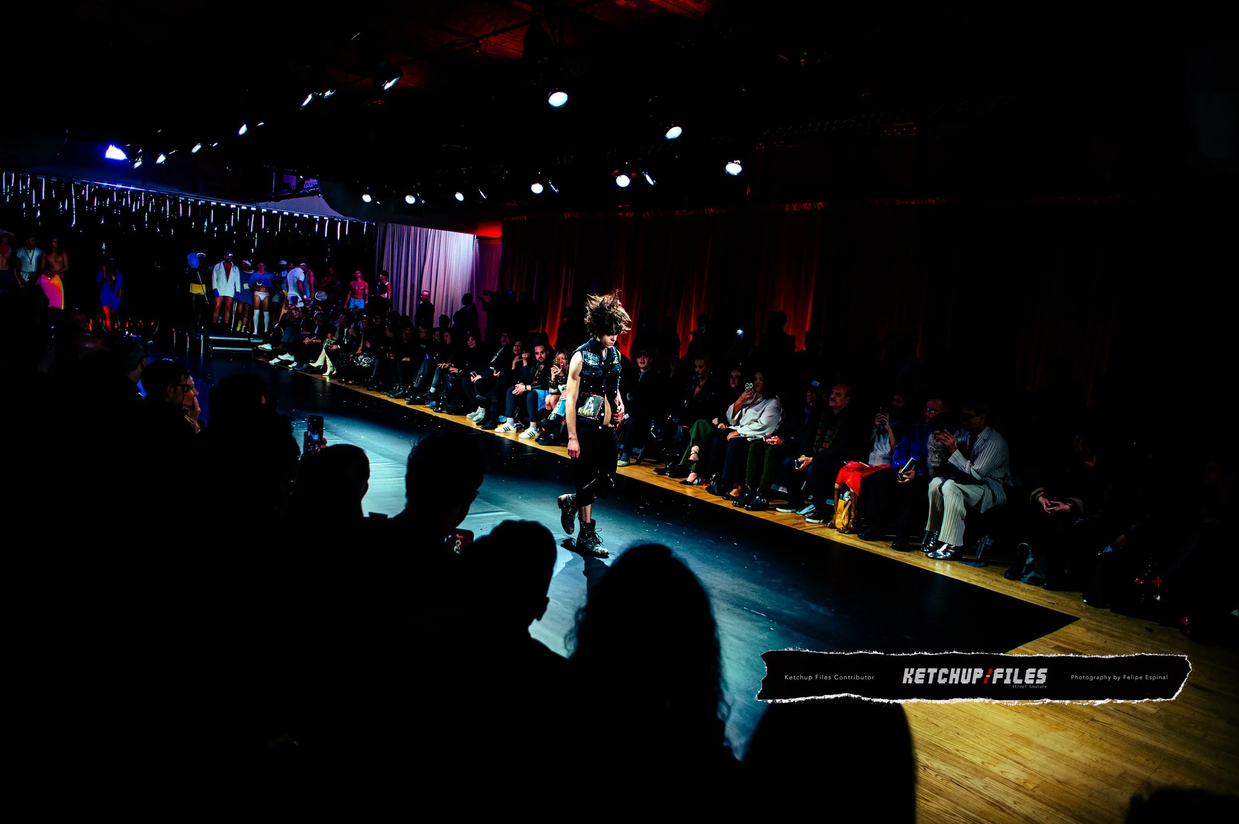 Fashion show runway with a model walking and audience seated on either side, dimly lit with red and blue lighting.